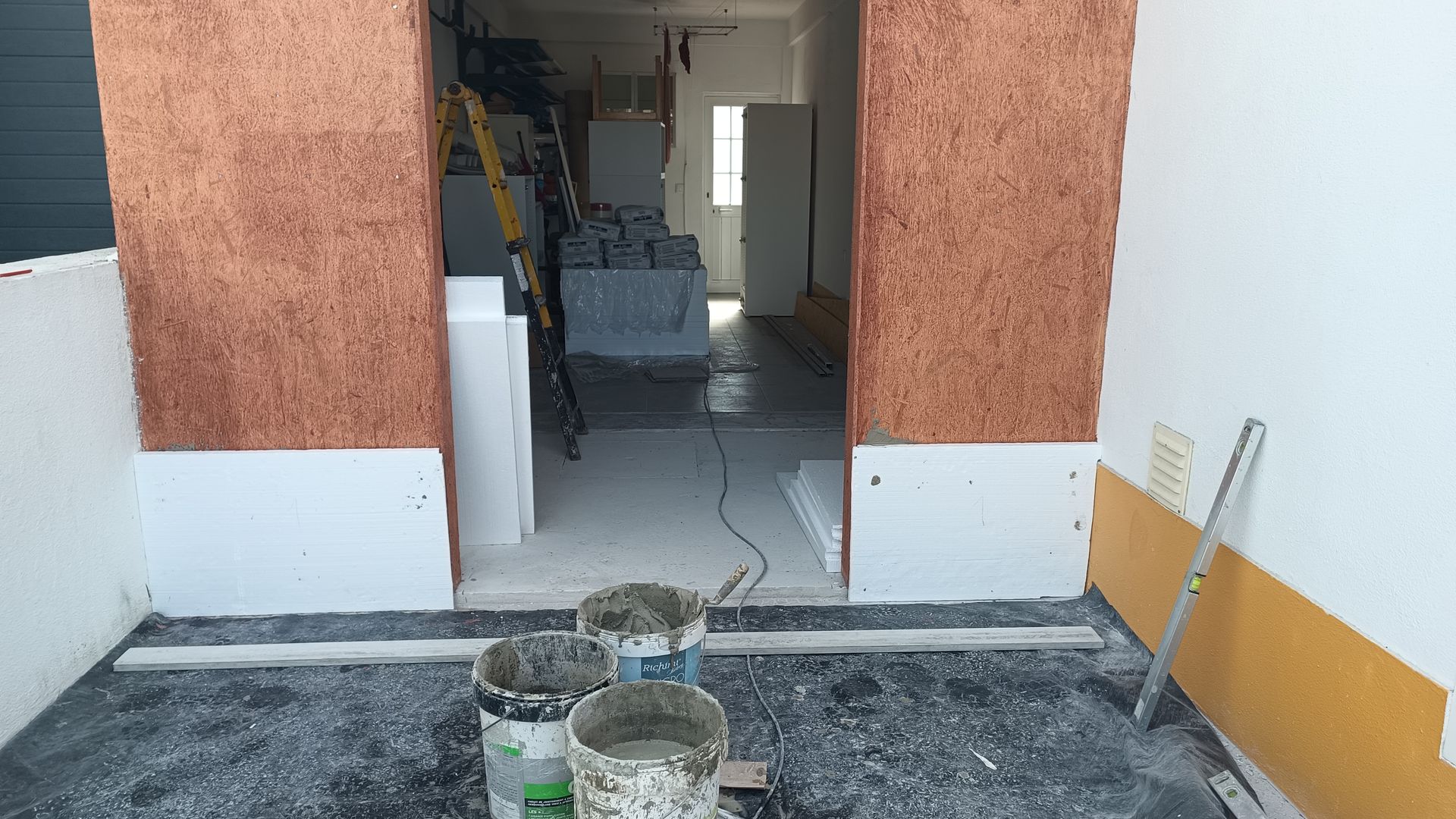 Entrada com colunas texturizadas em tom laranja, detalhes em branco e piso cinza. Baldes de material estão em primeiro plano.