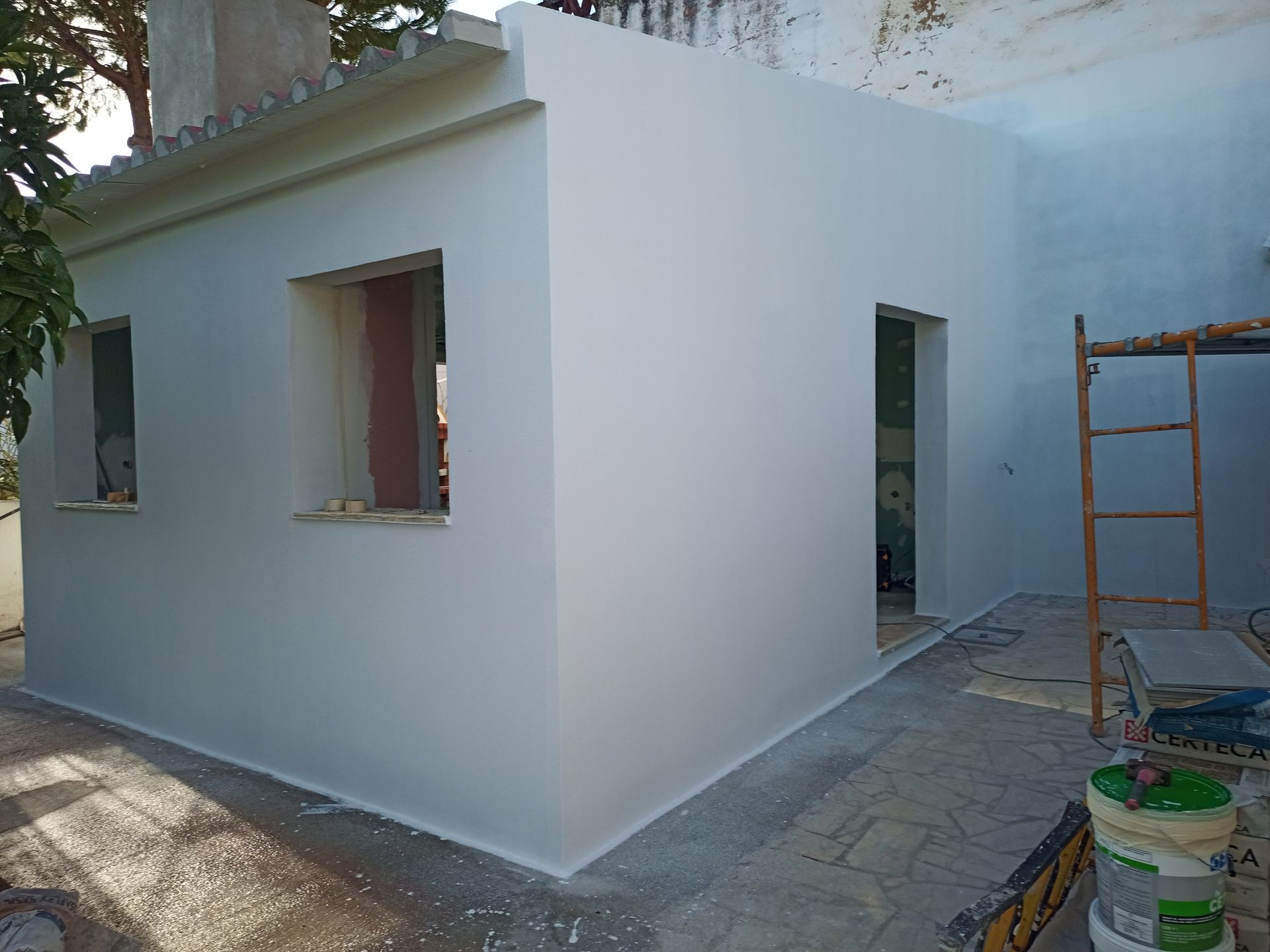 Exterior de um pequeno edifício branco com caixilhos de janelas e portas, em contexto de construção.