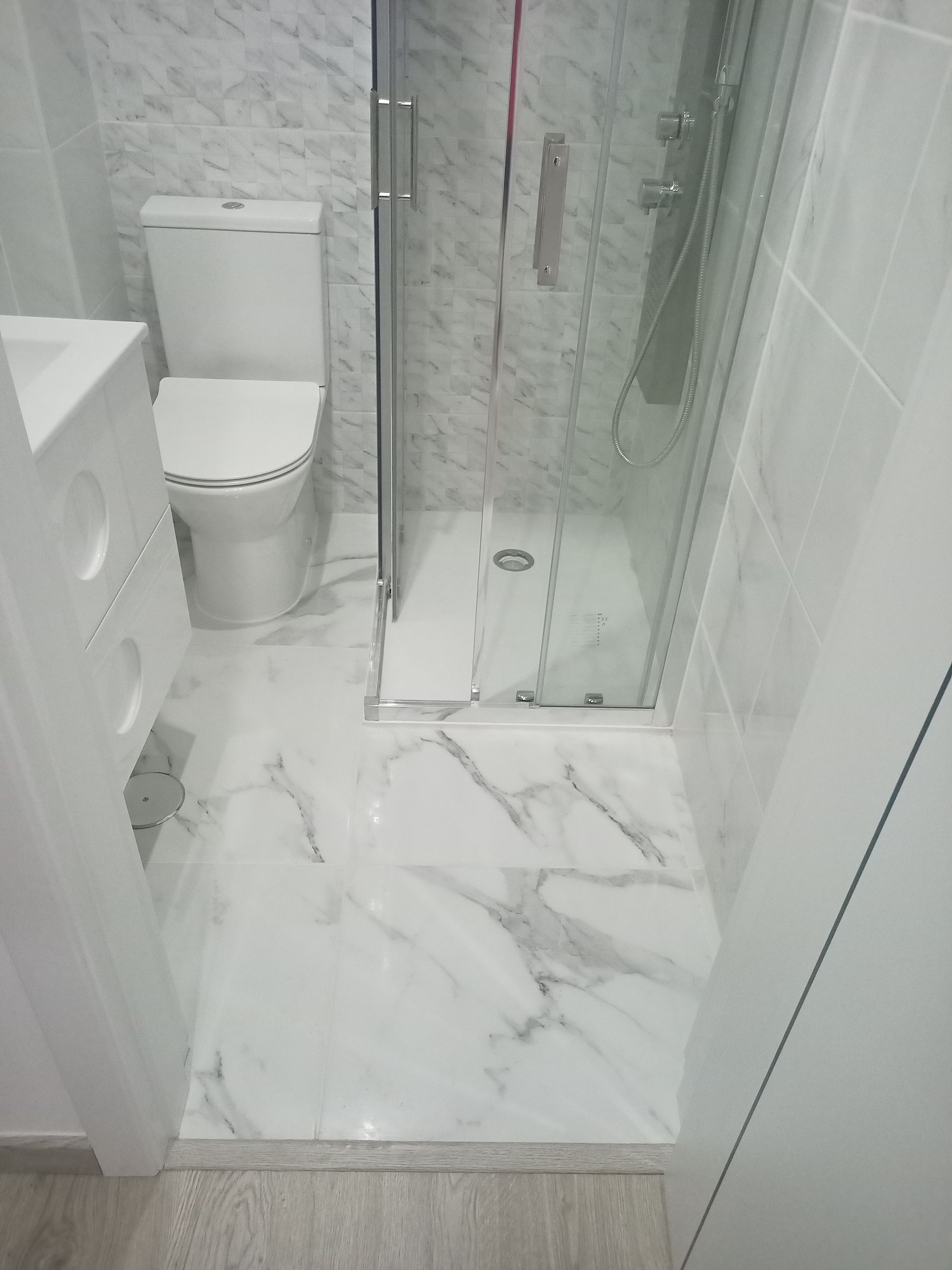 Um banheiro pequeno e moderno com azulejos que imitam mármore branco. Inclui vaso sanitário, chuveiro e pia.