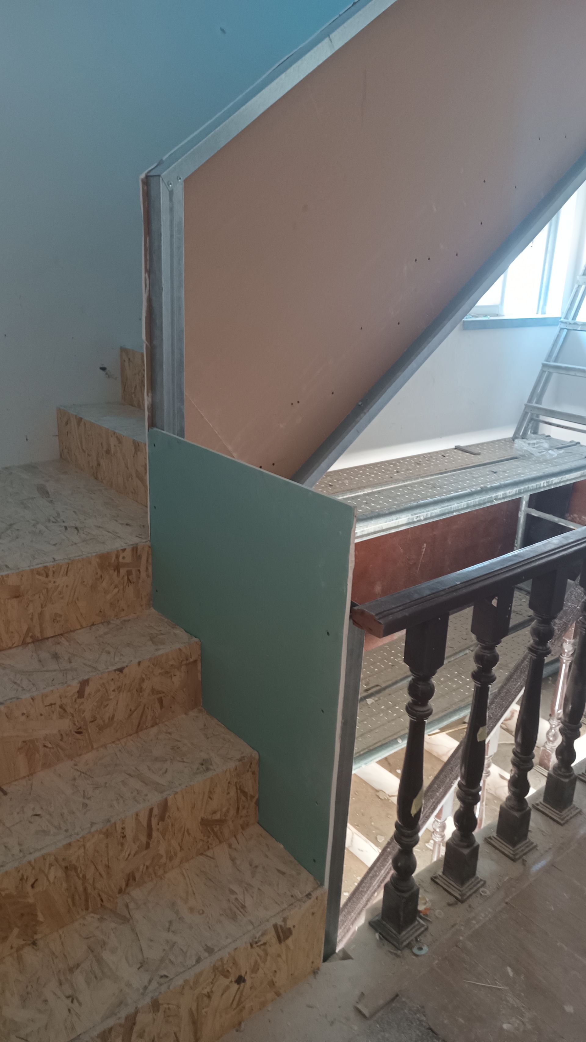 Escada em construção com drywall verde e bege, estrutura metálica e degraus de madeira com corrimão.