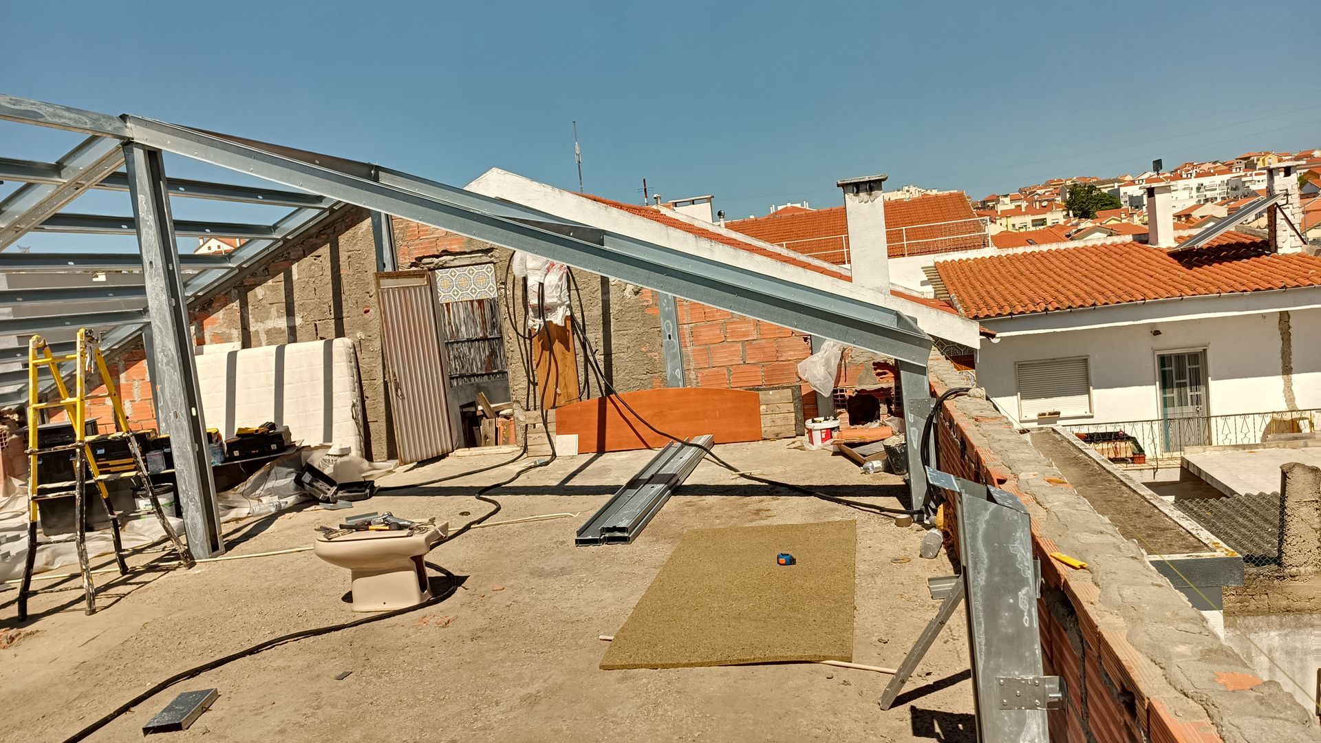 Estrutura metálica sobre um telhado, com prédios da cidade e céu azul ao fundo.