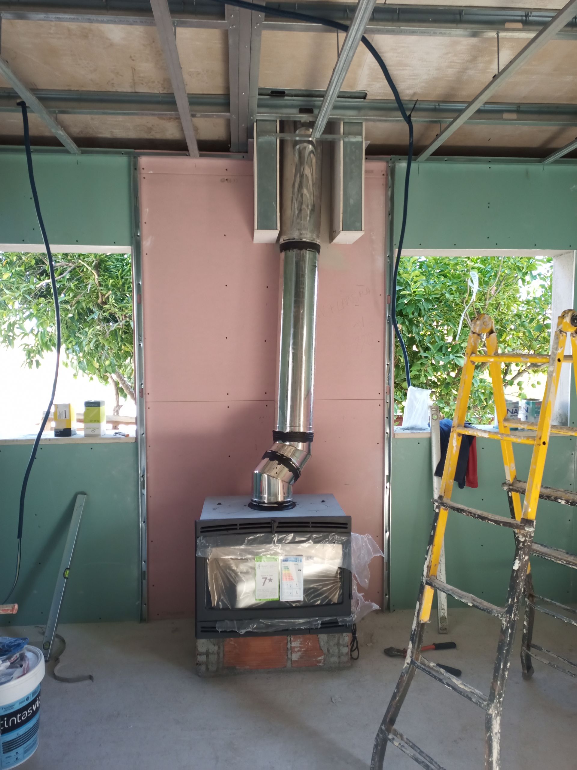 Instalação de fogão a lenha em um cômodo em construção; o tubo de chaminé metálico atravessa o teto.