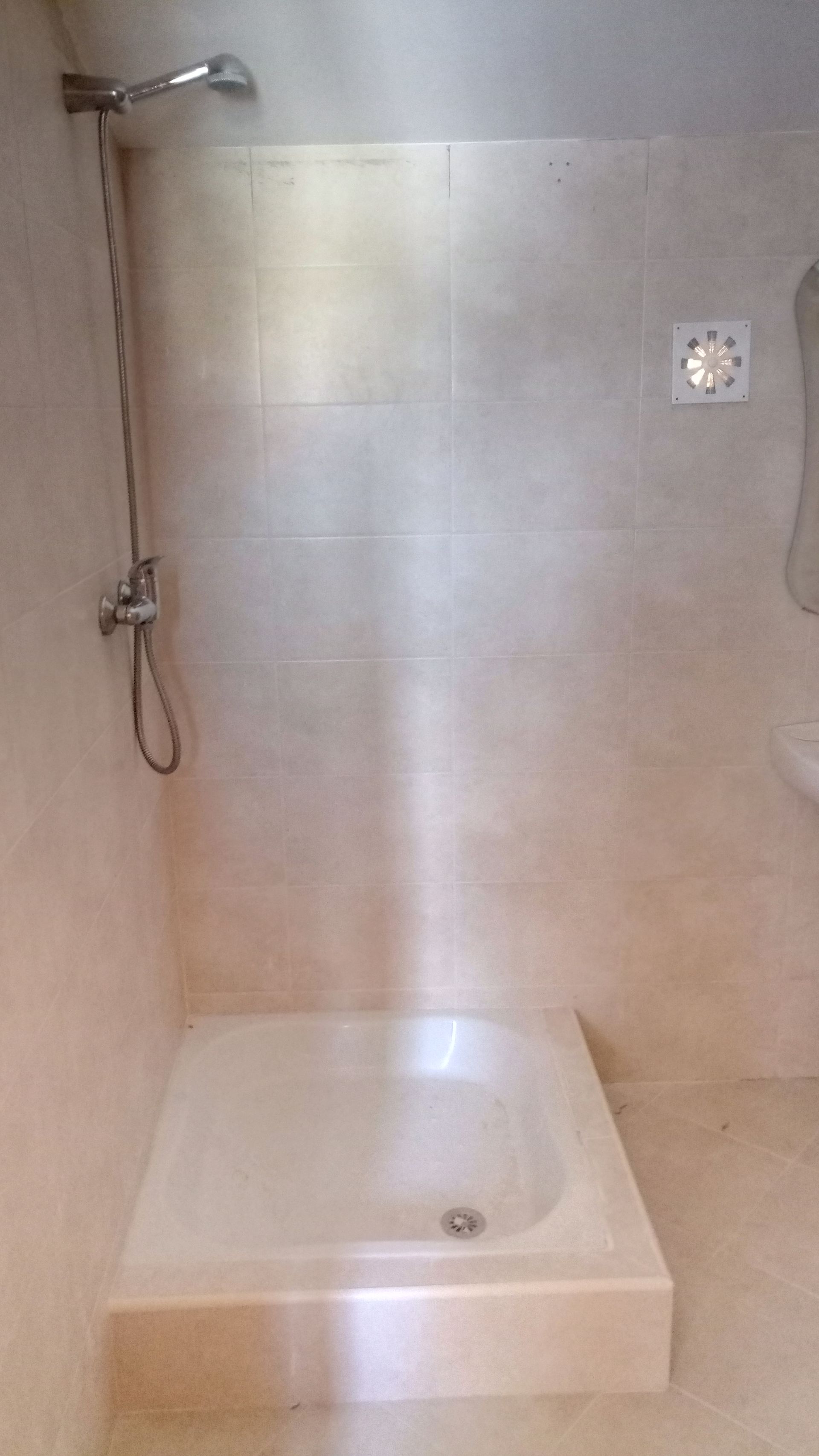 Chuveiro com base branca e paredes revestidas com azulejos. Chuveiro e torneira à esquerda, pequena saída de ar quadrada à direita.