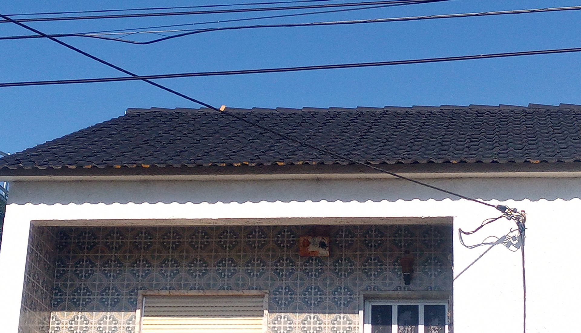 Fachada branca da casa com telhado de telhas e fiação elétrica suspensa.