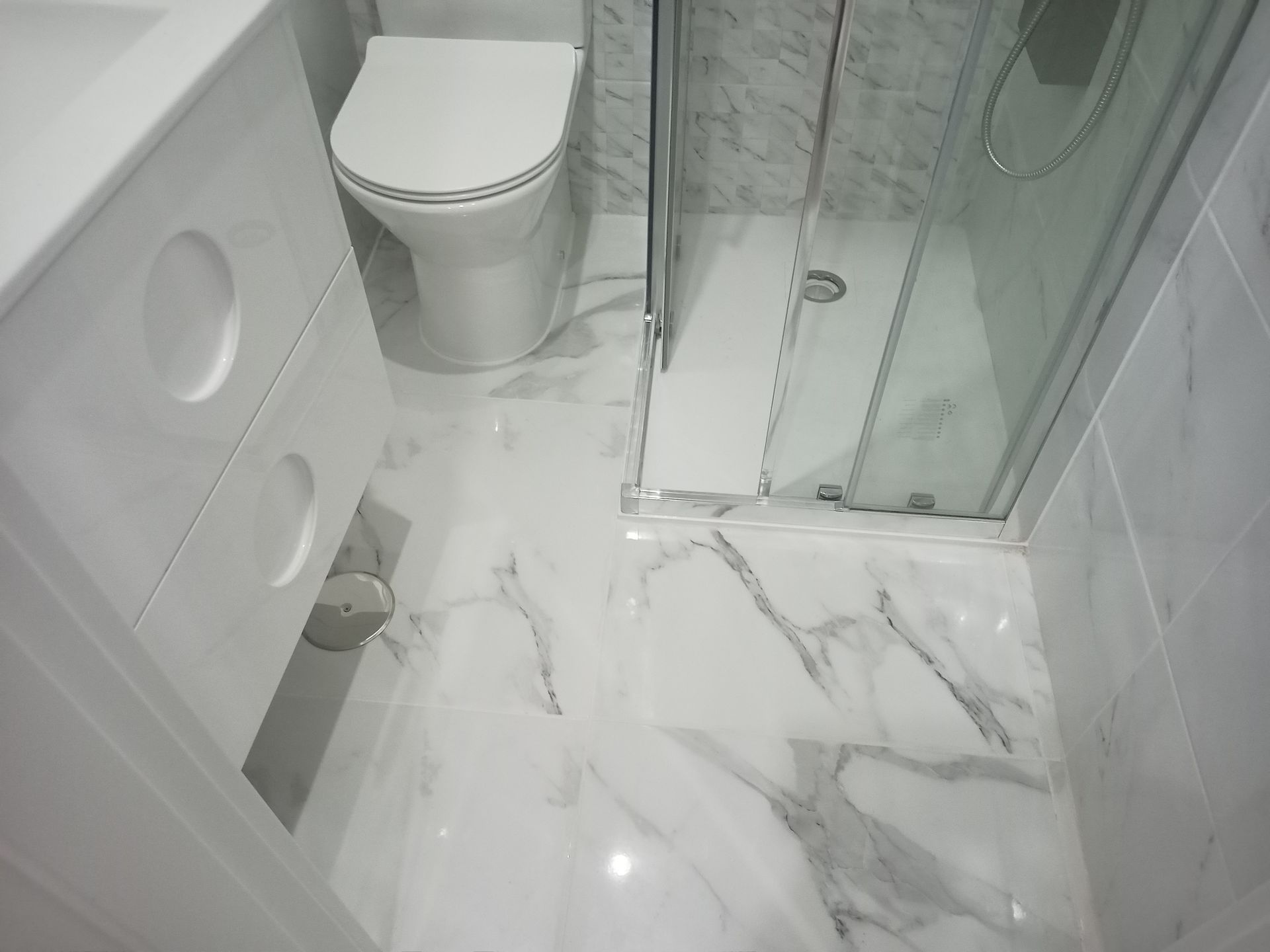 Banheiro branco com piso de azulejo imitando mármore, vaso sanitário, chuveiro e pia.