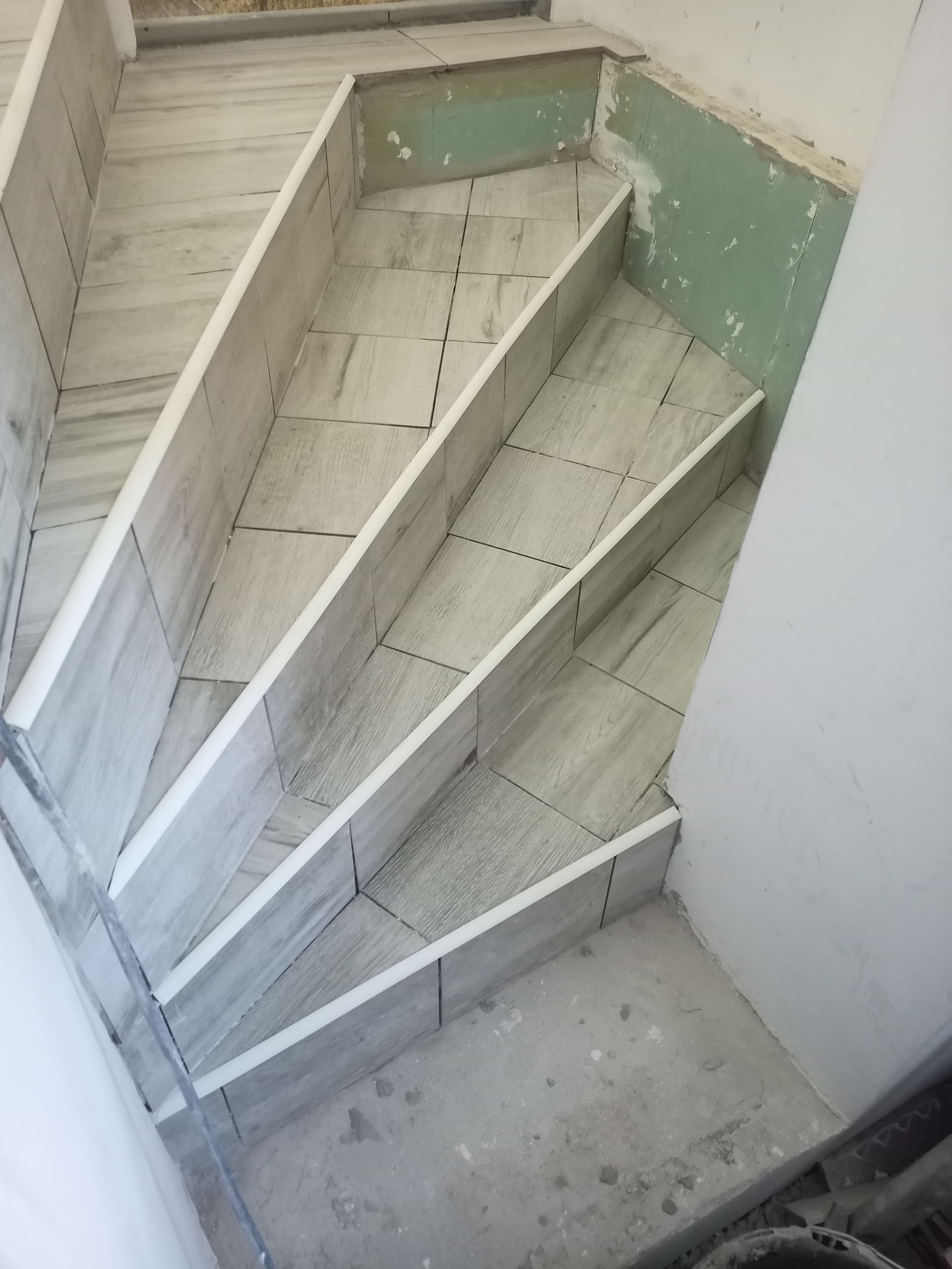 Escada em construção com azulejos claros e concreto cinza. É possível ver gesso acartonado verde na parede.
