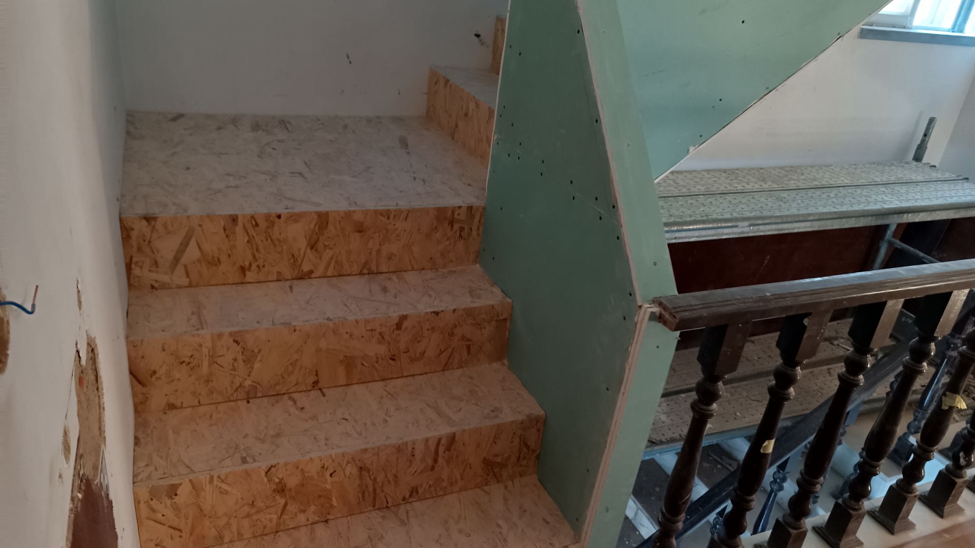 Escada com degraus em OSB (painel de madeira orientada) e uma parede verde, que leva a um segundo andar com corrimão de madeira.