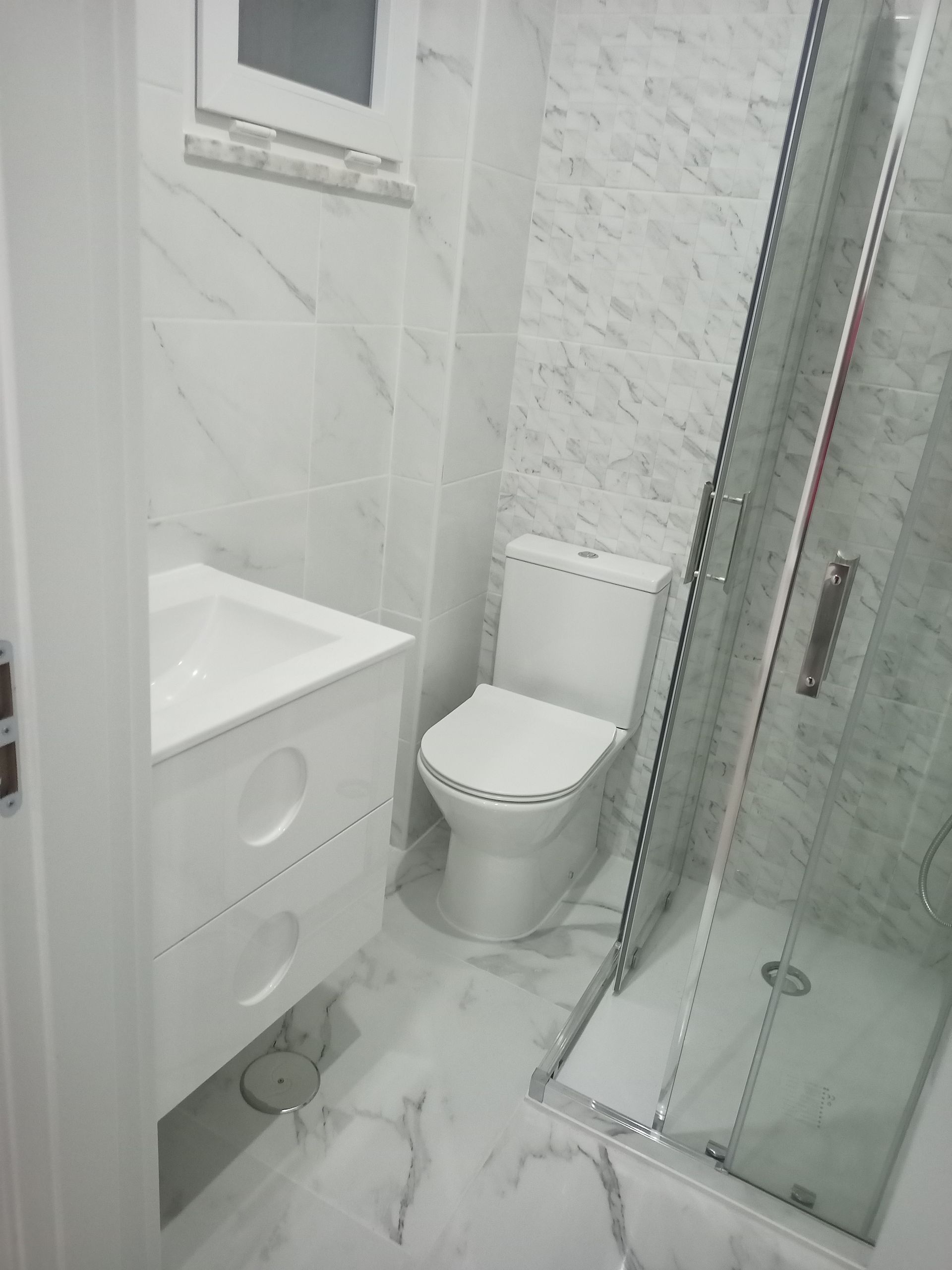 Banheiro branco com vaso sanitário, pia e chuveiro. Azulejos com estampa de mármore revestem as paredes e o piso.