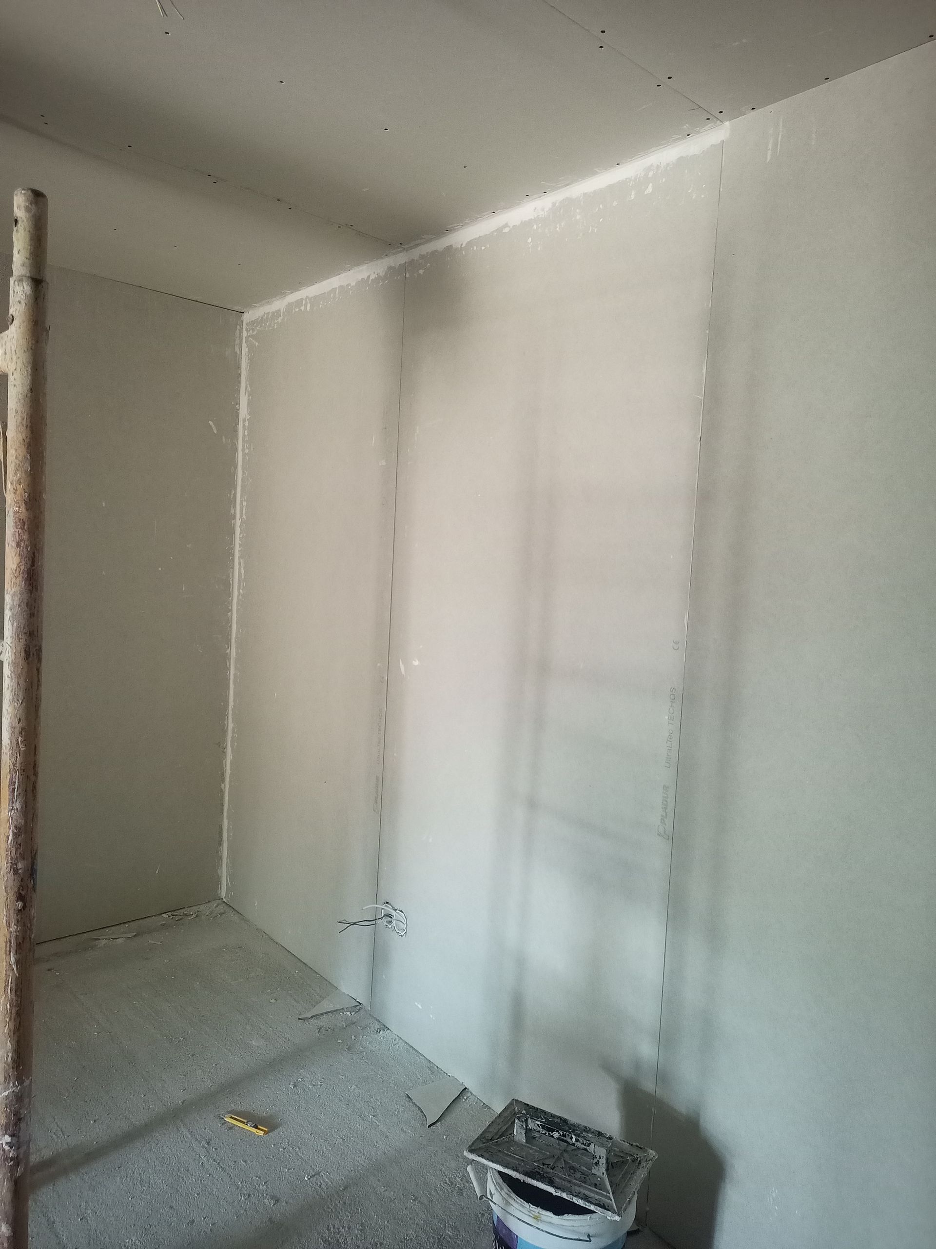 Interior de uma sala em construção com paredes rebocadas de cinza e piso de concreto. Uma escada de madeira é visível.