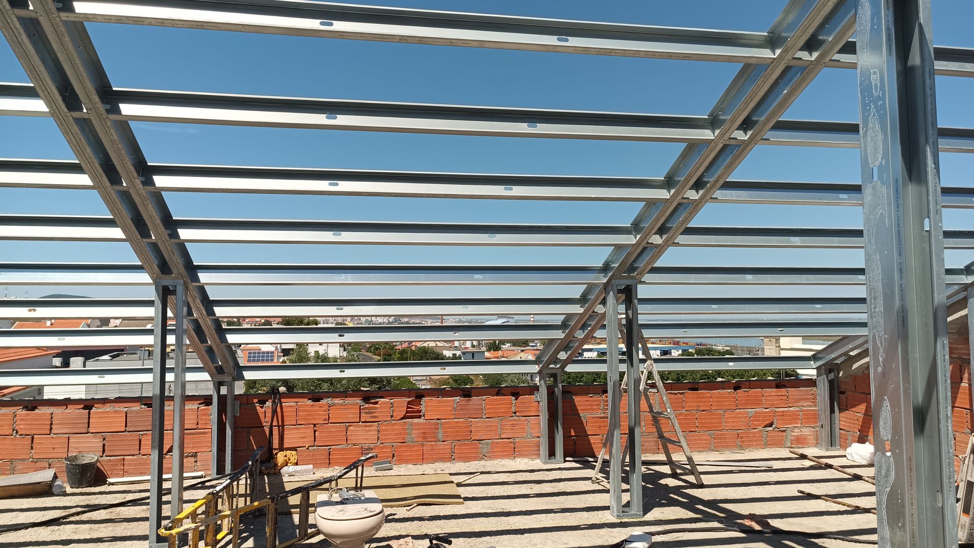 Estrutura metálica para um telhado em construção, tendo como pano de fundo uma parede de tijolos vermelhos e um céu azul claro.