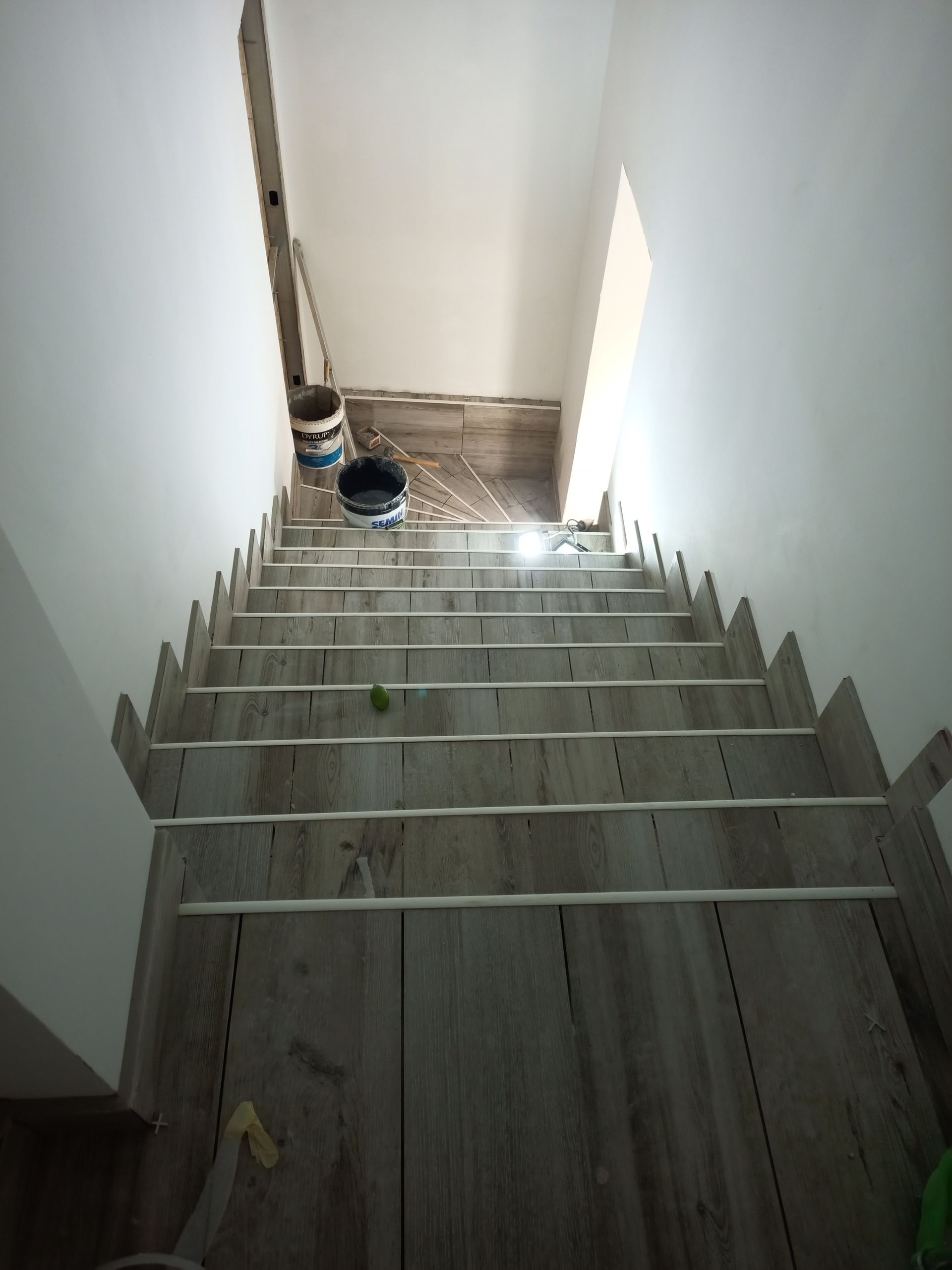 Escada em construção; degraus revestidos com azulejos imitando madeira e com espaçadores. Parte superior da escada iluminada por luz natural.