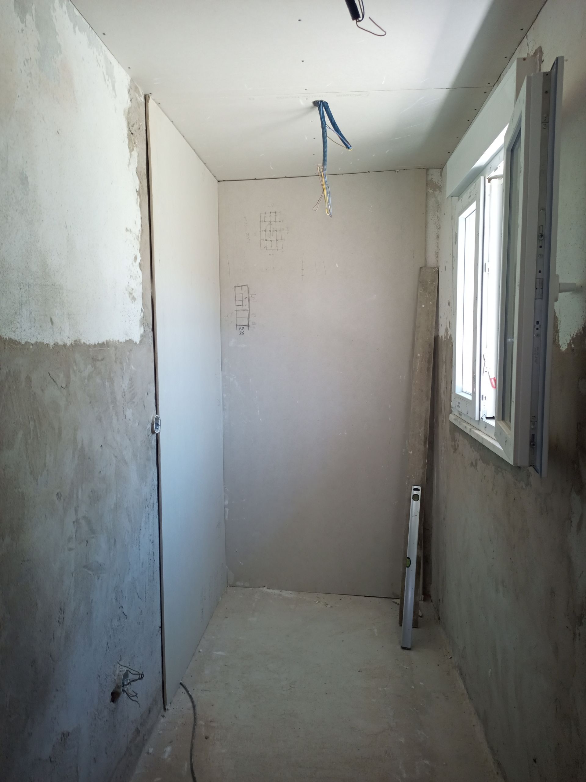 Interior de um pequeno cômodo em construção, com paredes de gesso acartonado e uma janela.