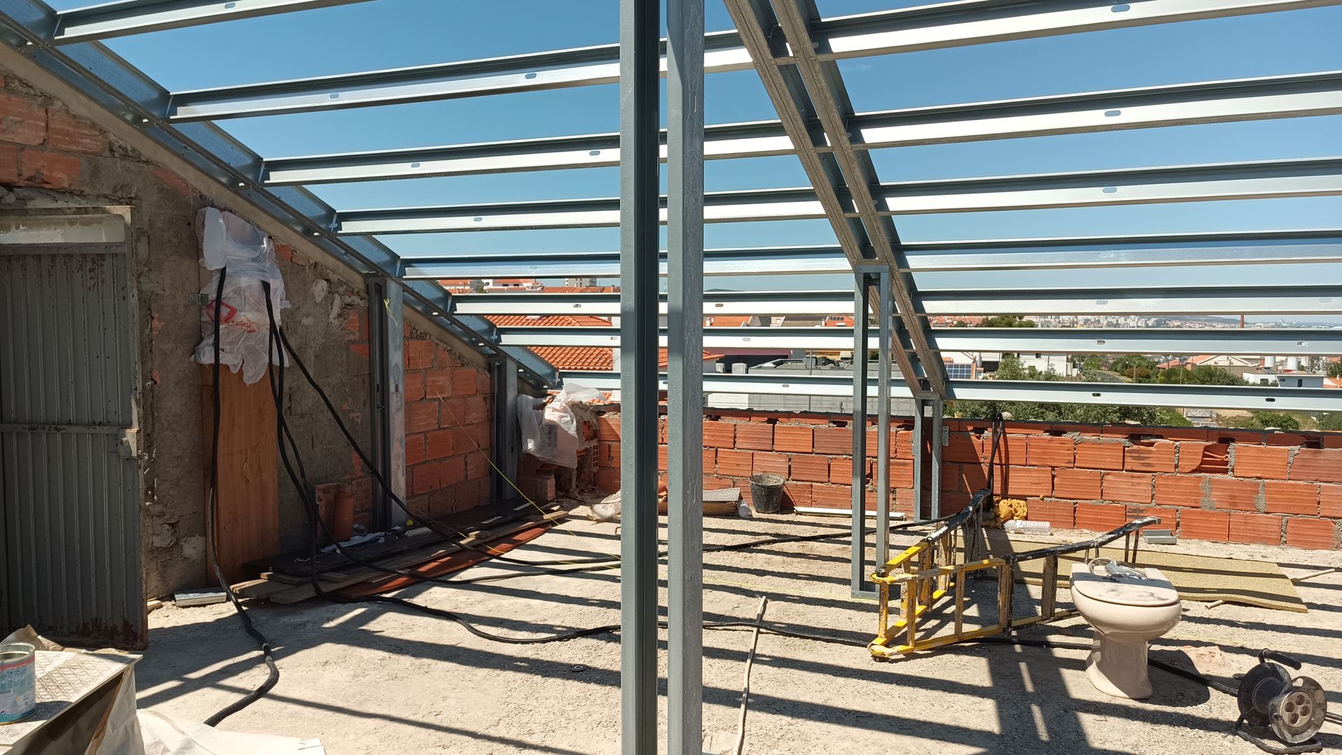 Canteiro de obras com vigas metálicas para o telhado, paredes de tijolos à vista, iluminação natural.