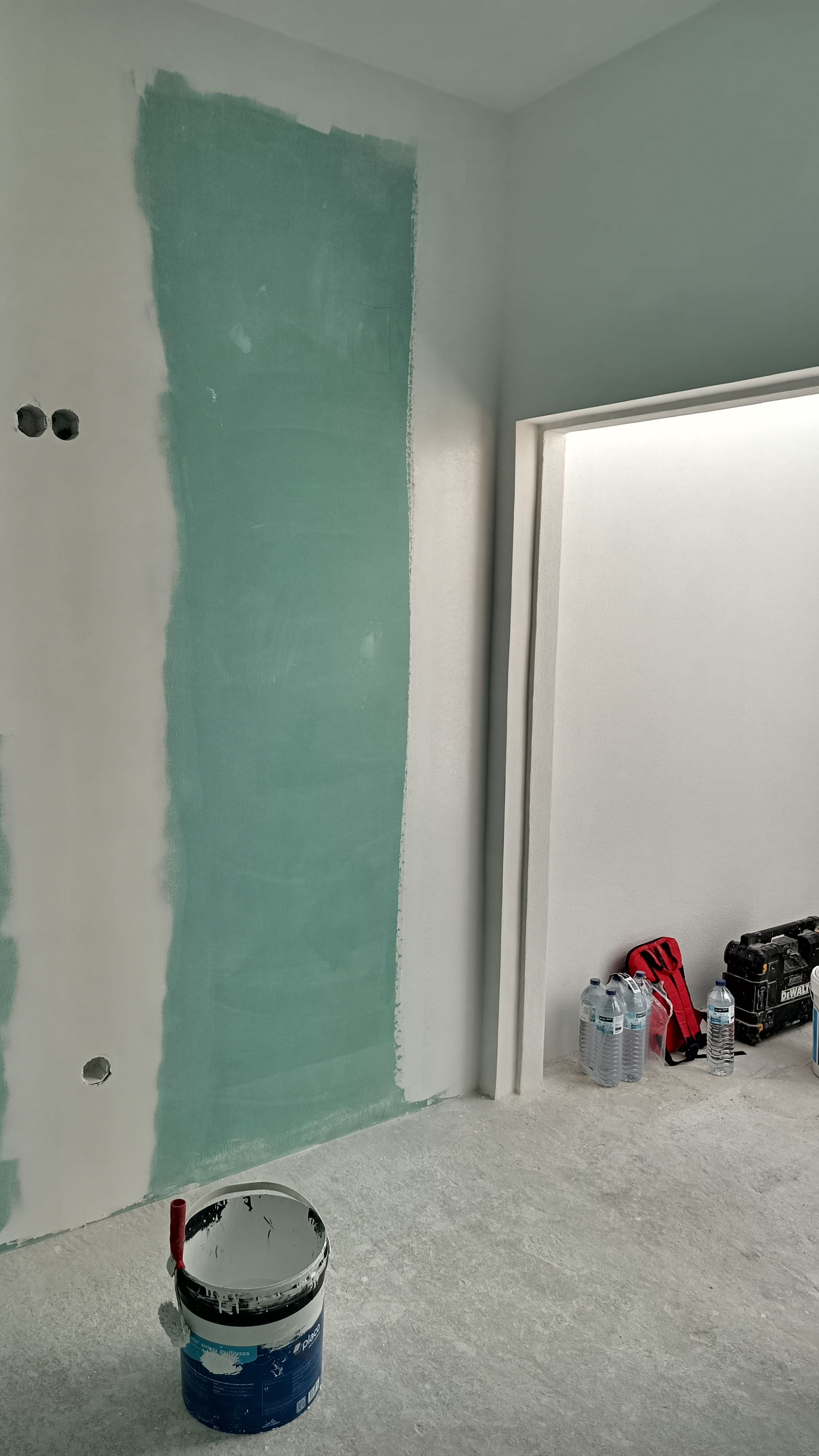Interior de um cômodo com paredes de gesso inacabadas; uma parede é verde, a porta e um balde de tinta são visíveis.