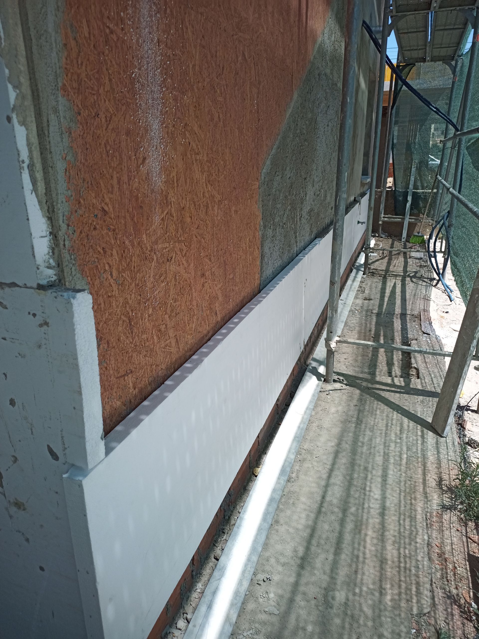 Canteiro de obras: parede externa mostrando camadas de material em tons de marrom, verde e branco. Andaimes visíveis.