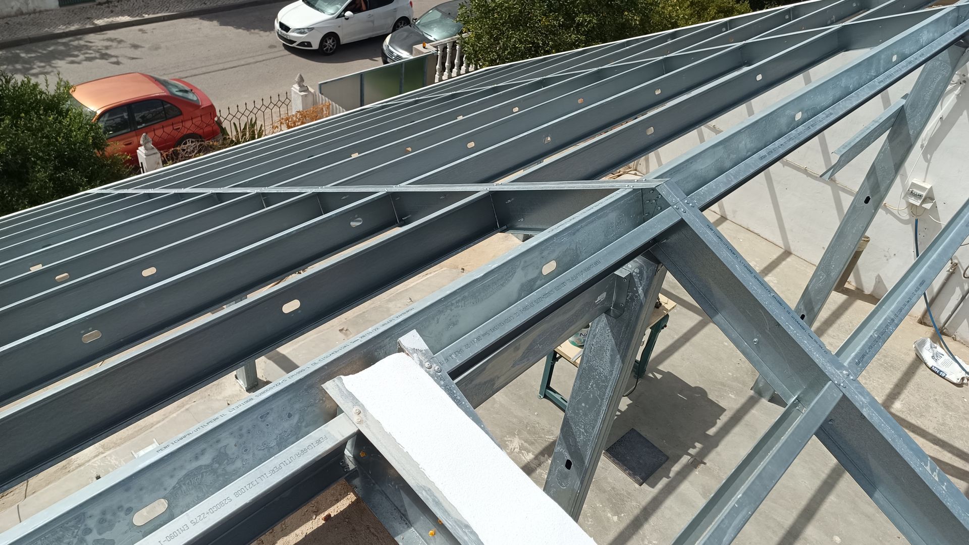 Estrutura metálica do telhado, vista externa. Vigas e barras de metal cinza formam a estrutura do telhado, com veículos e árvores ao fundo.