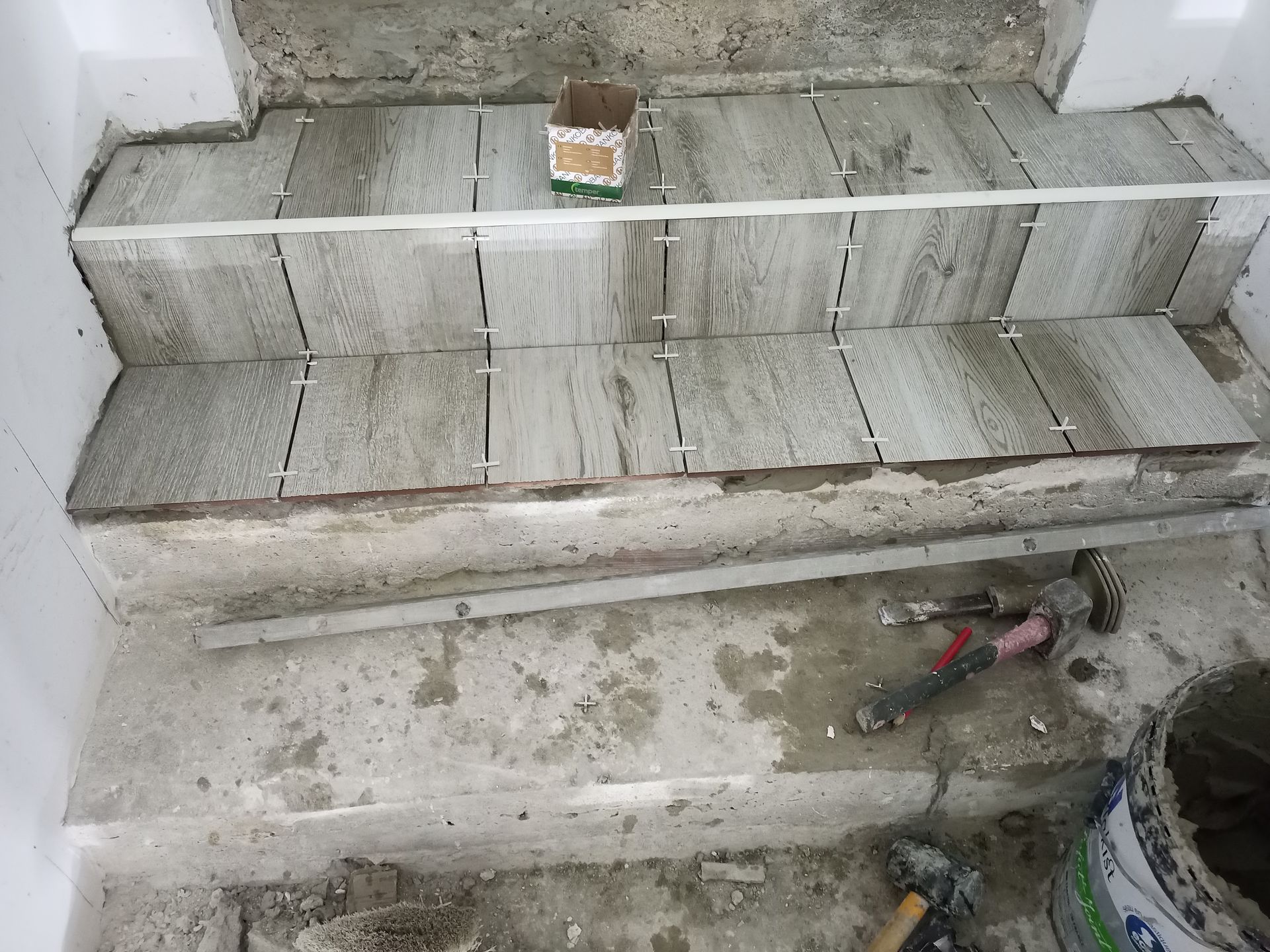 Escada em construção com ladrilhos cinza, espaçadores e ferramentas. Paredes de concreto e brancas são visíveis.