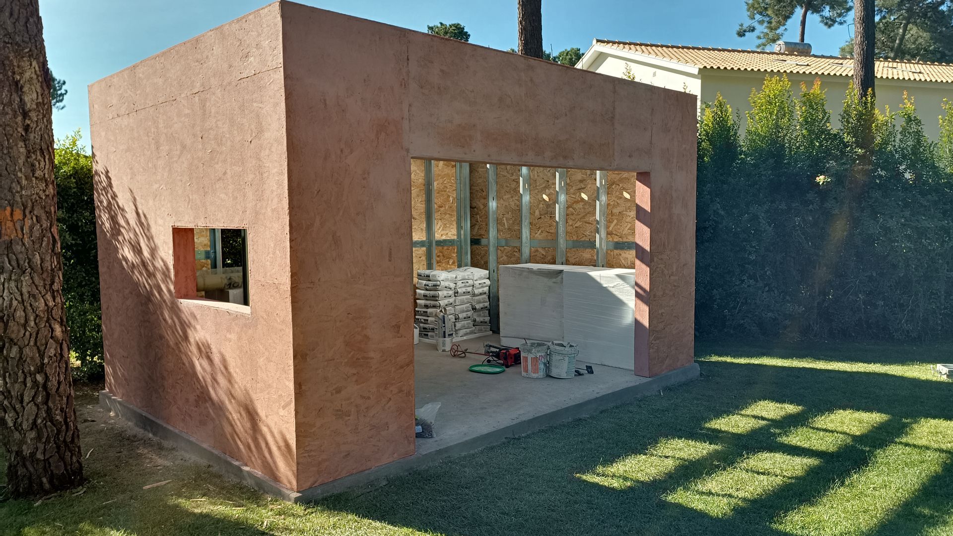 Construção de um pequeno galpão marrom-avermelhado em um quintal gramado. Portas e janelas abertas.