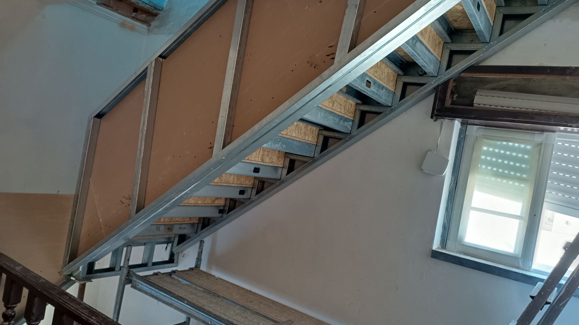 Escada em construção: estrutura metálica, madeira, paredes inacabadas, janela e corrimão de madeira marrom.