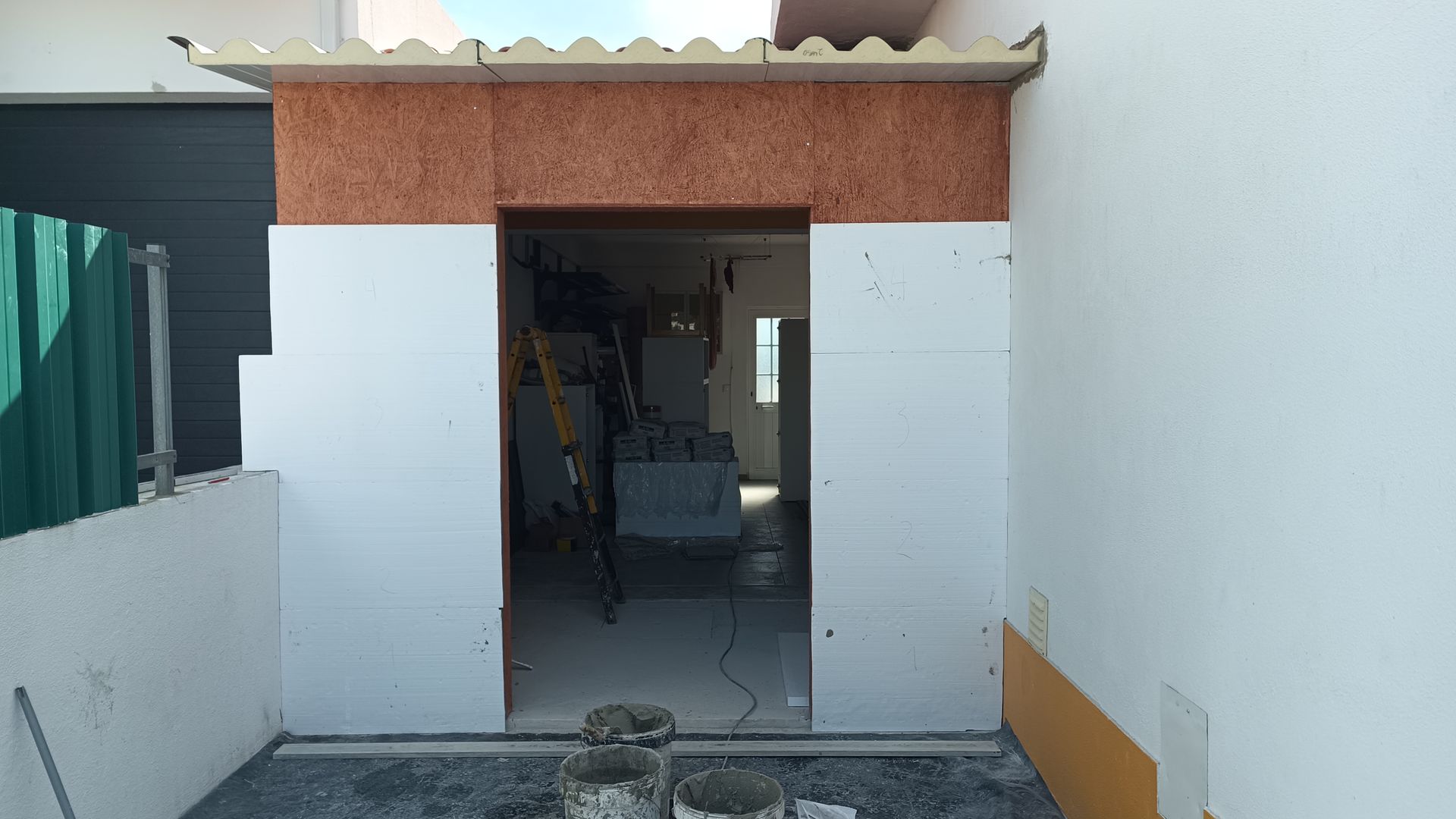 Porta externa emoldurada por painéis brancos com detalhes em marrom-avermelhado na parte superior, dando acesso a uma sala com materiais de construção.