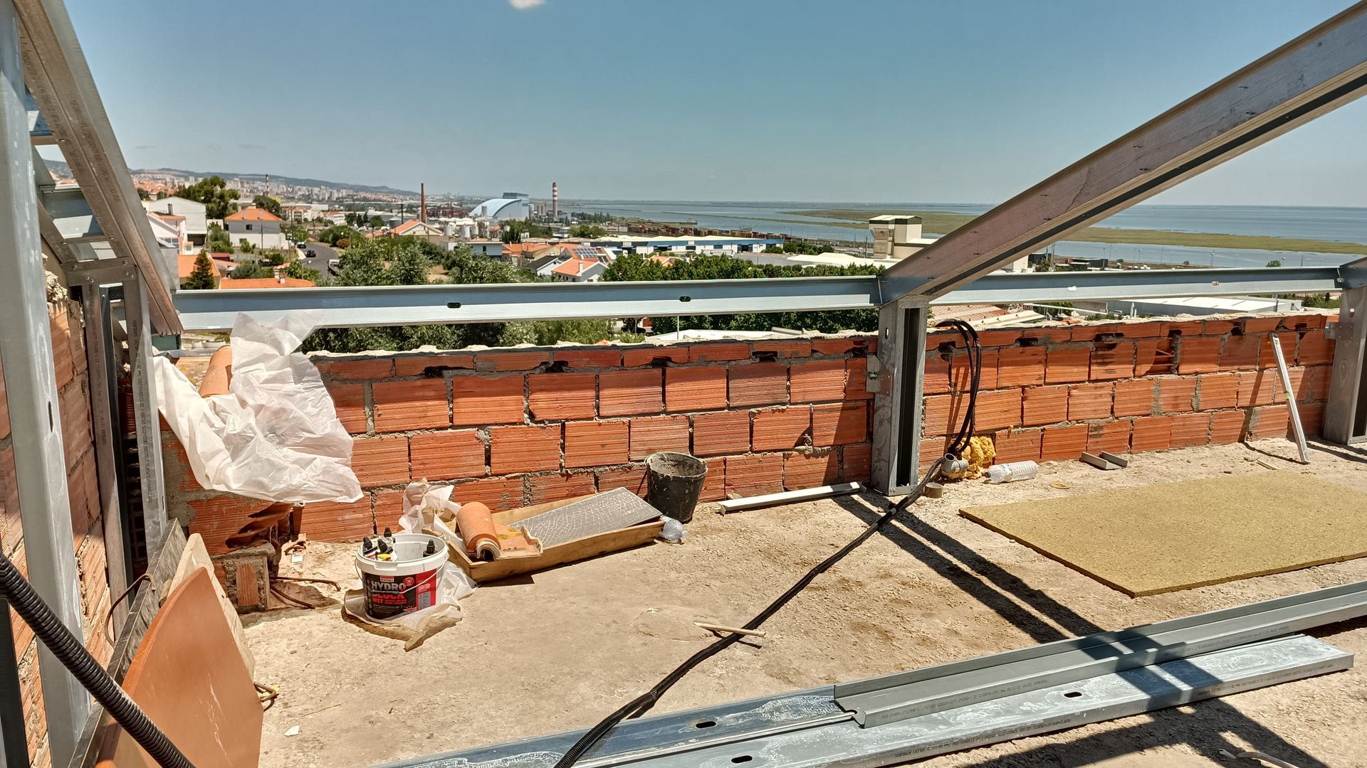 Canteiro de obras com parede de tijolos vermelhos, estrutura metálica e paisagem urbana visível ao fundo.