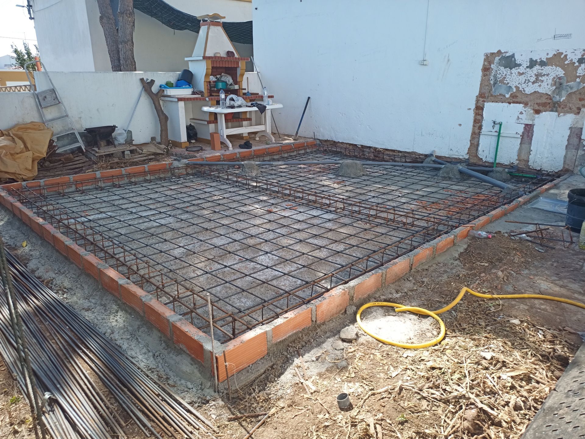 Canteiro de obras com estrutura de fundação em concreto armado, malha de vergalhões e tijolos ao redor.