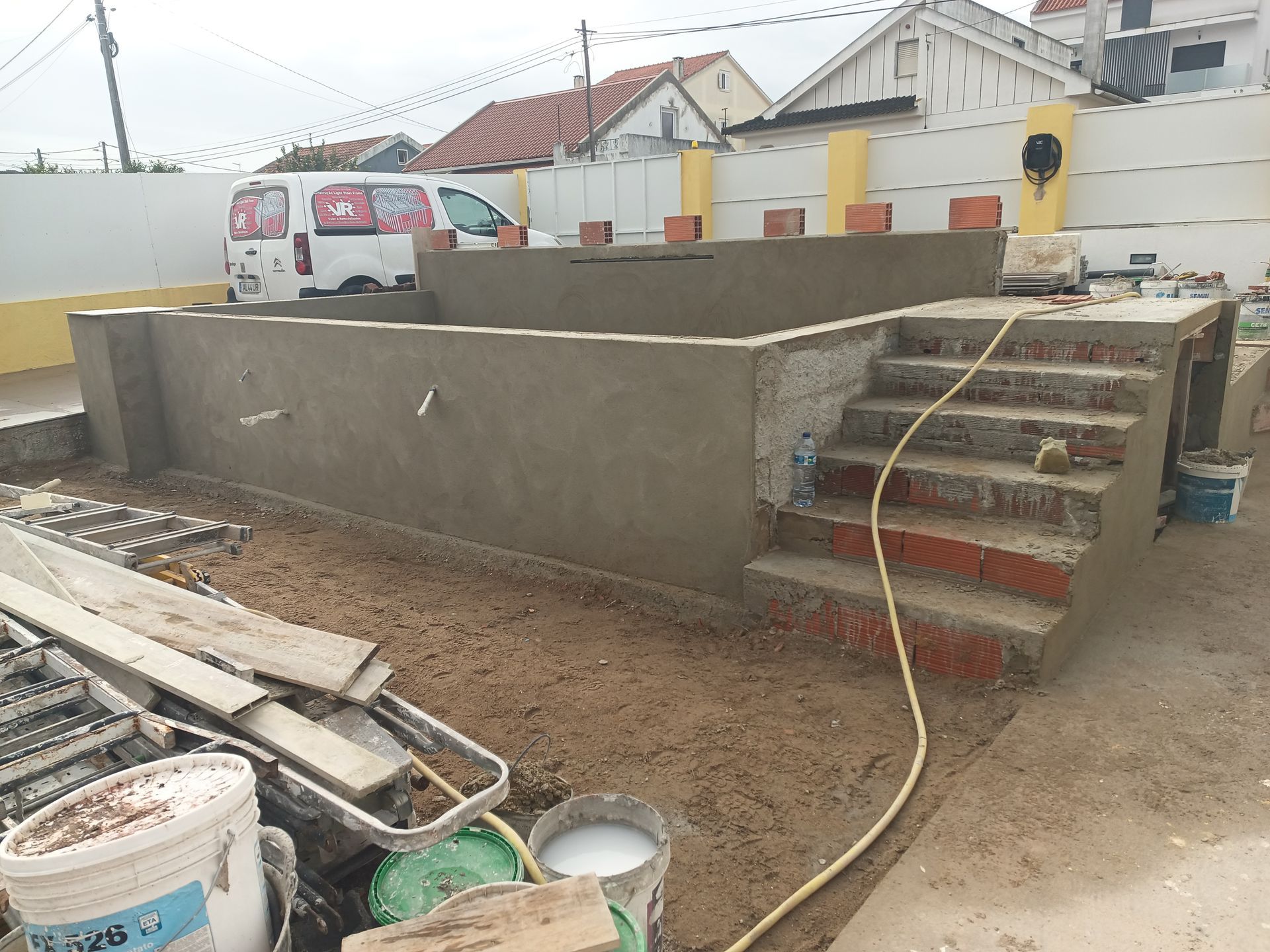 Construção de uma estrutura de concreto com degraus, cercada por materiais de construção e uma van branca ao fundo.