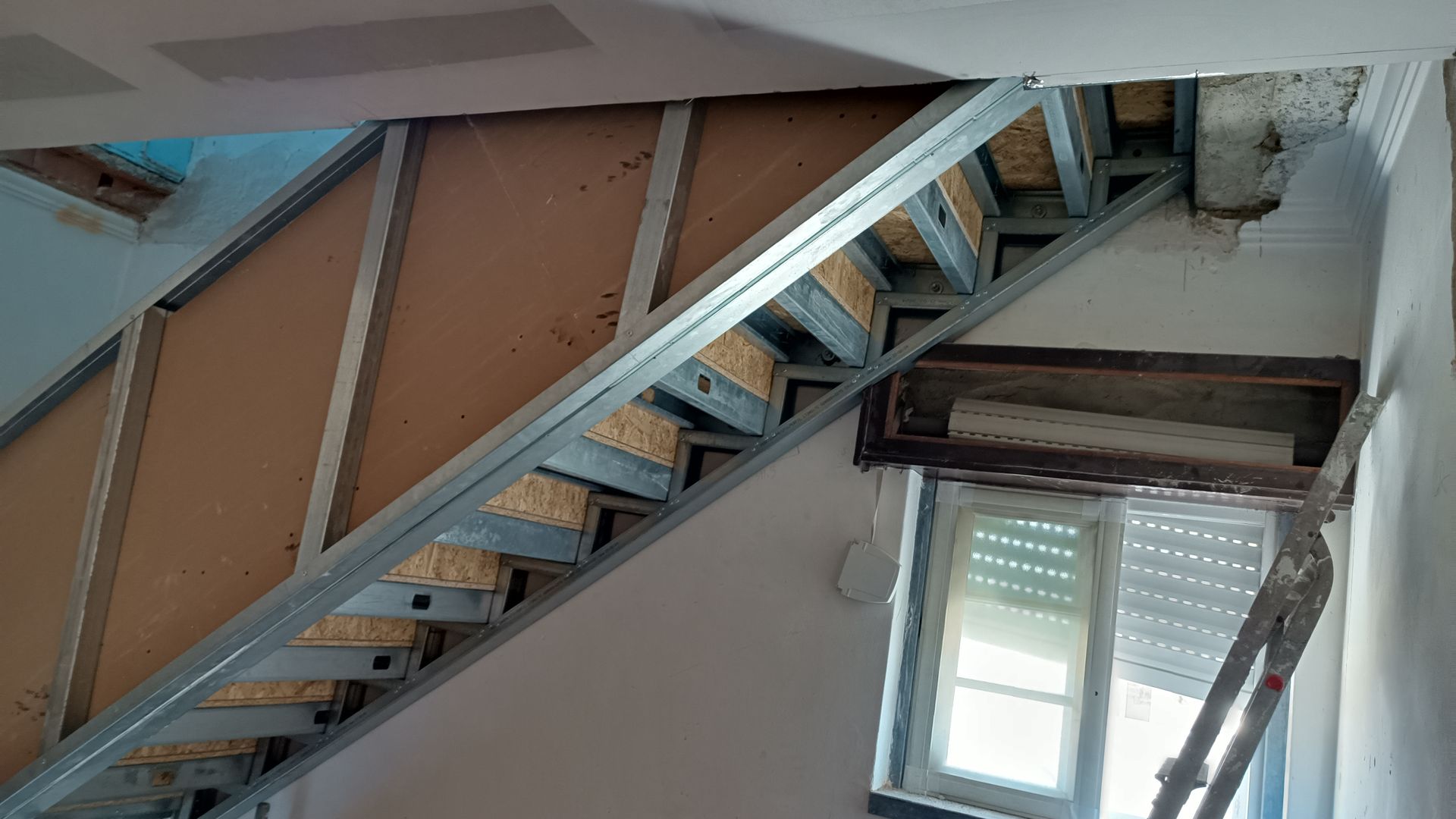 Estrutura metálica da escada com degraus de madeira instalados, visível contra uma parede e uma janela.