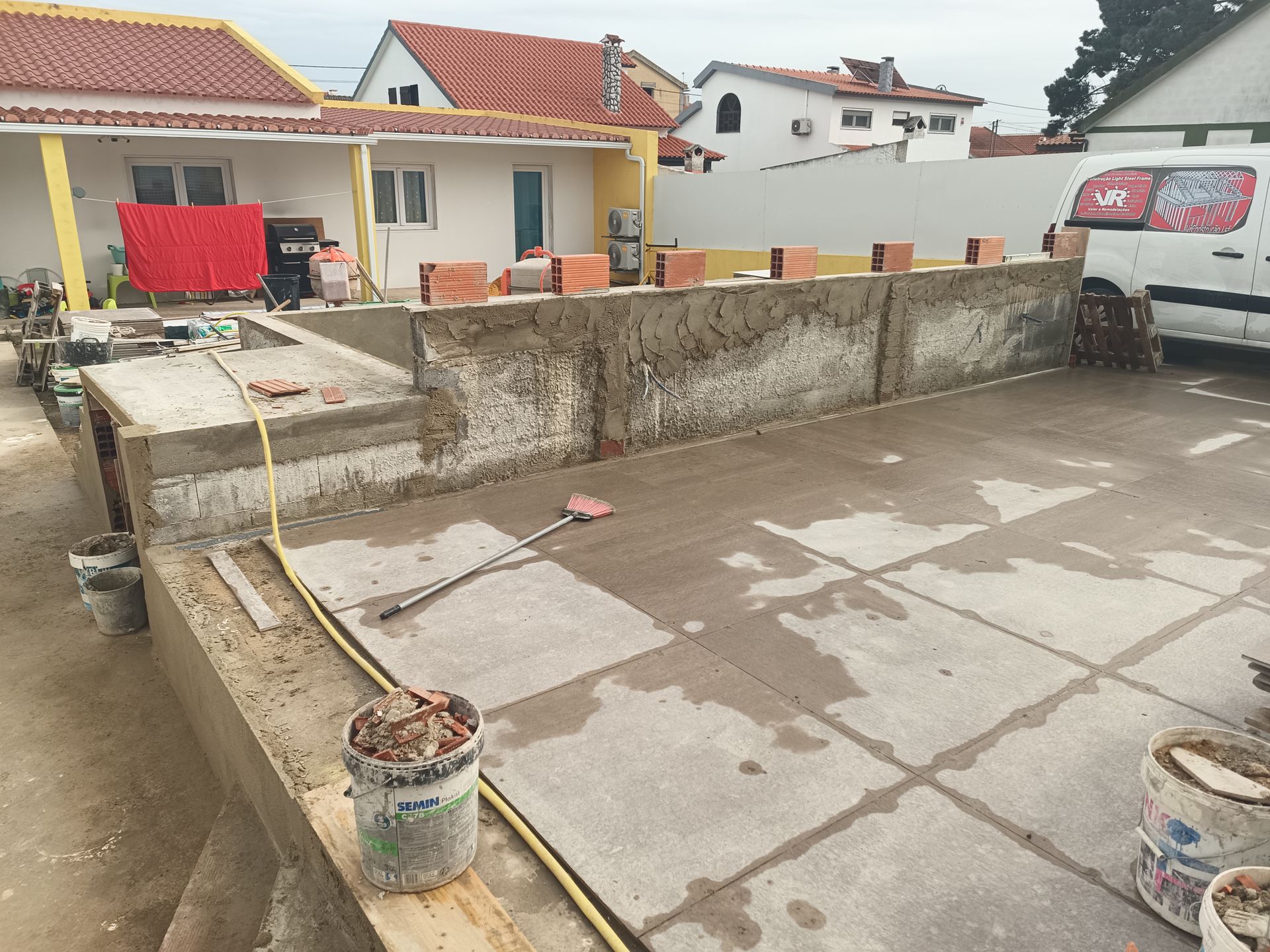 Canteiro de obras: estrutura de concreto com tampo de tijolos, mangueira amarela, baldes e uma van branca.