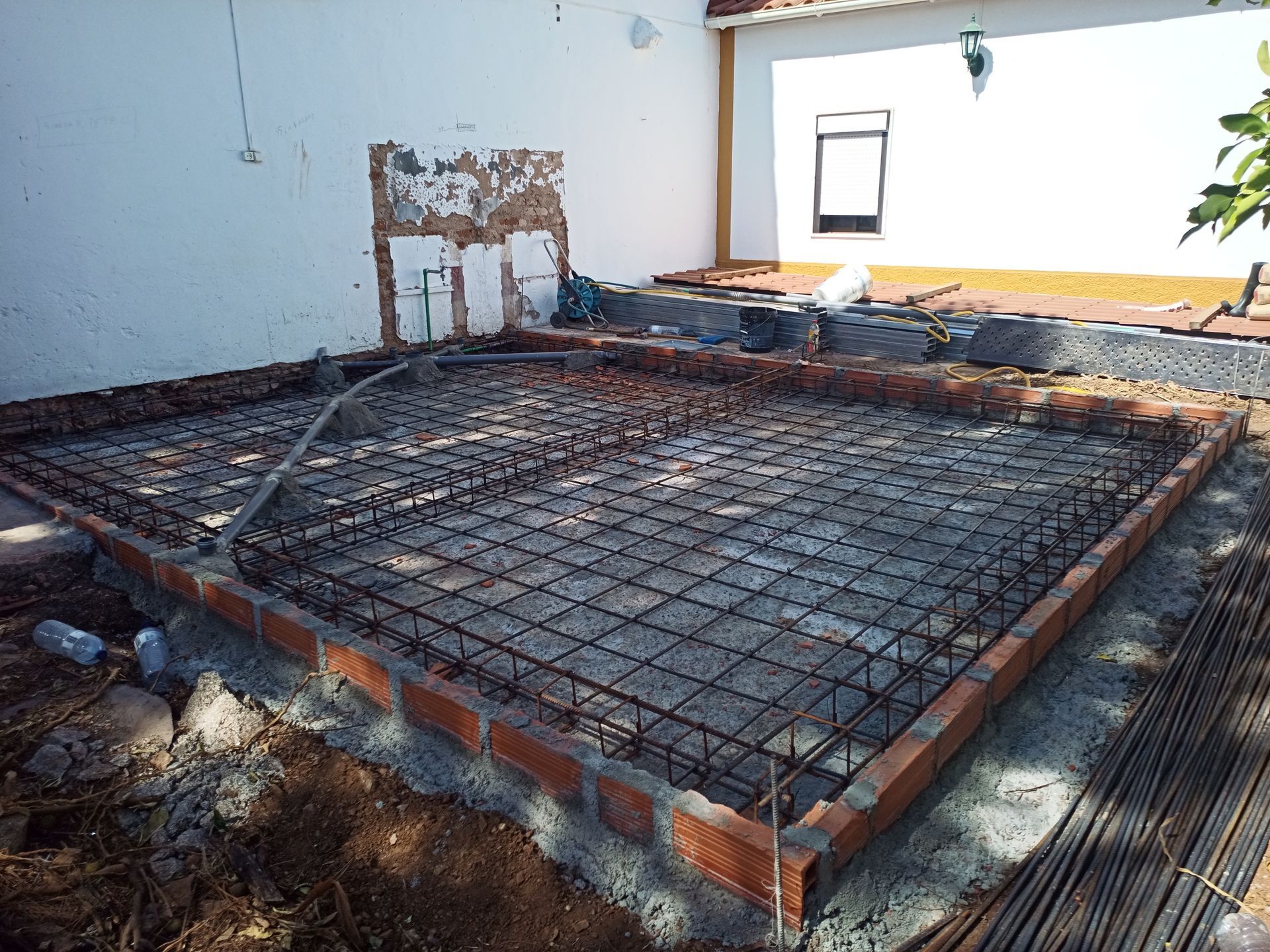 Canteiro de obras: estrutura perimetral de tijolos com malha de vergalhões para laje de concreto; parede branca ao fundo.