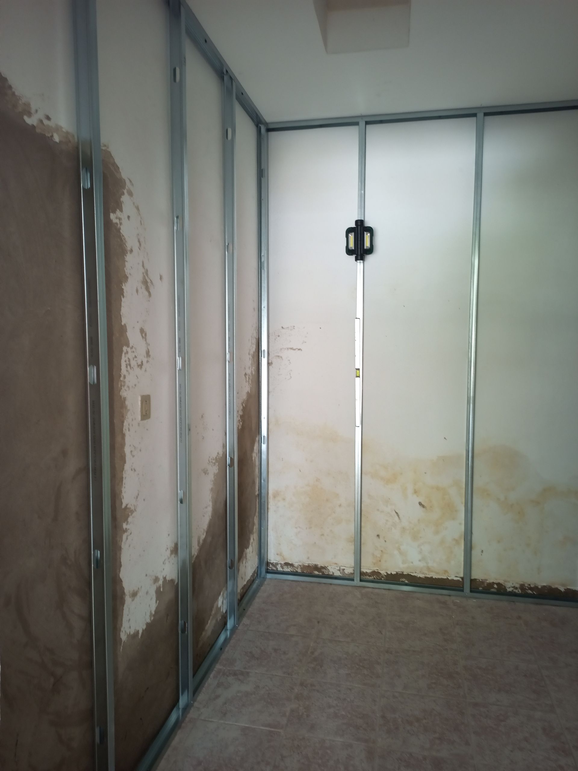 Interior de um cômodo em construção; perfis metálicos sustentam as paredes, que apresentam danos causados ​​pela água. Manchas marrons são visíveis.