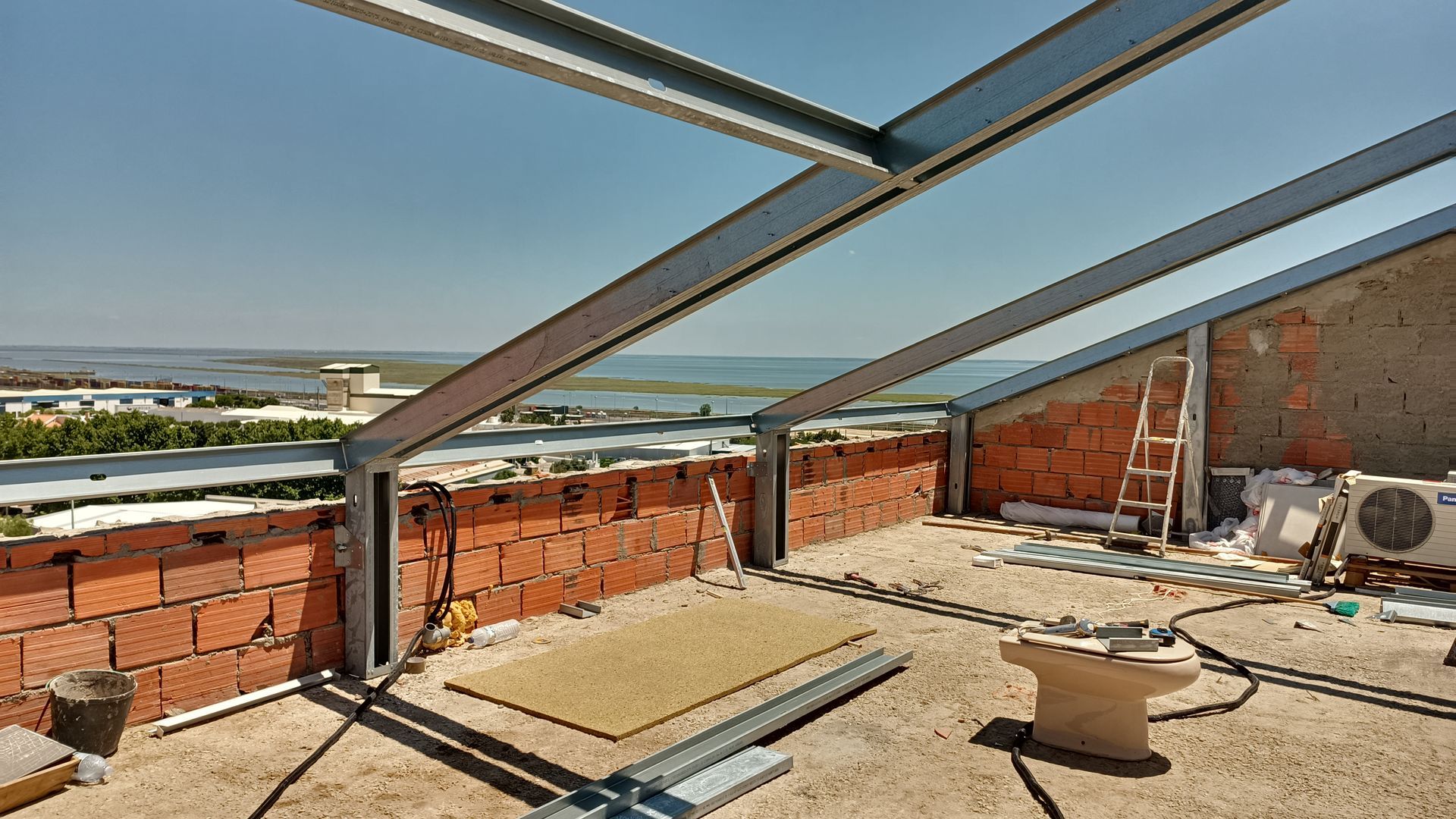 Canteiro de obras com vigas de aço, paredes de tijolos e vista para a água.