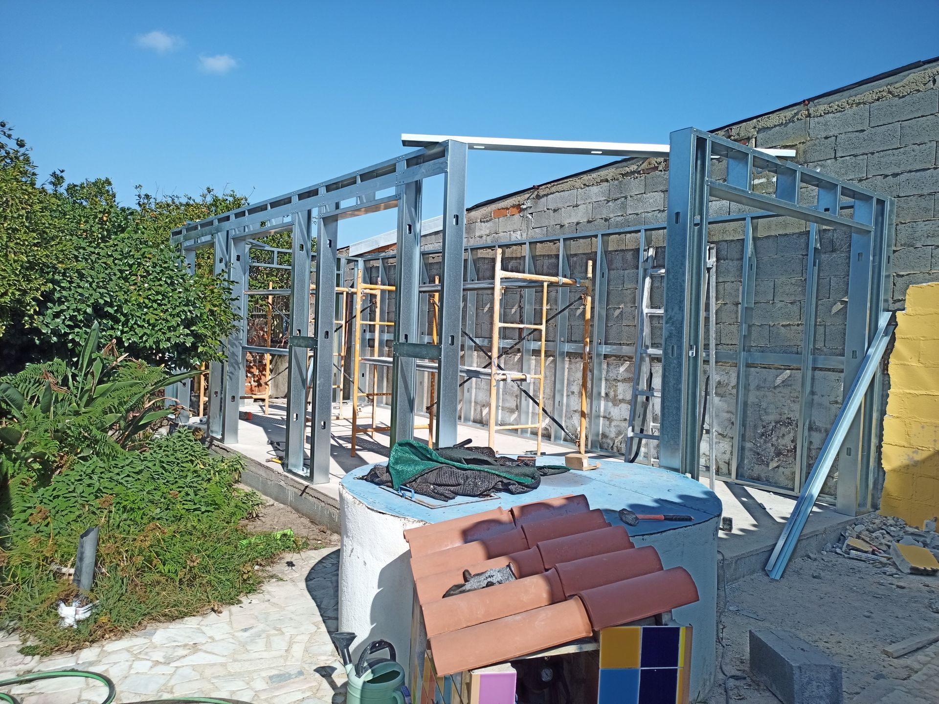 Estrutura metálica de um edifício em construção, encostada a uma parede de tijolos, com telhas e materiais de cobertura no chão.