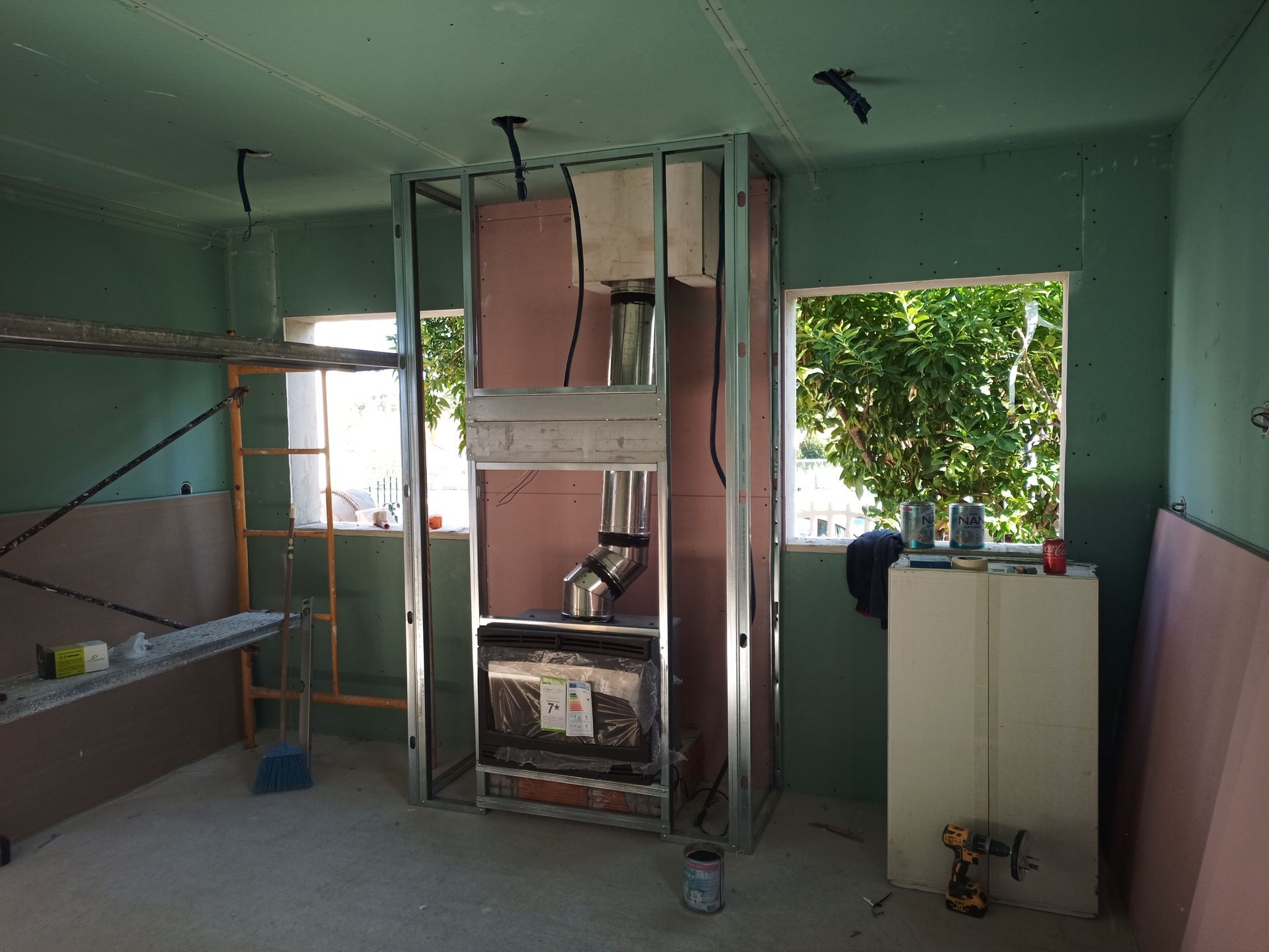 Sala em construção com estrutura de lareira em metal, drywall, janelas e andaimes.