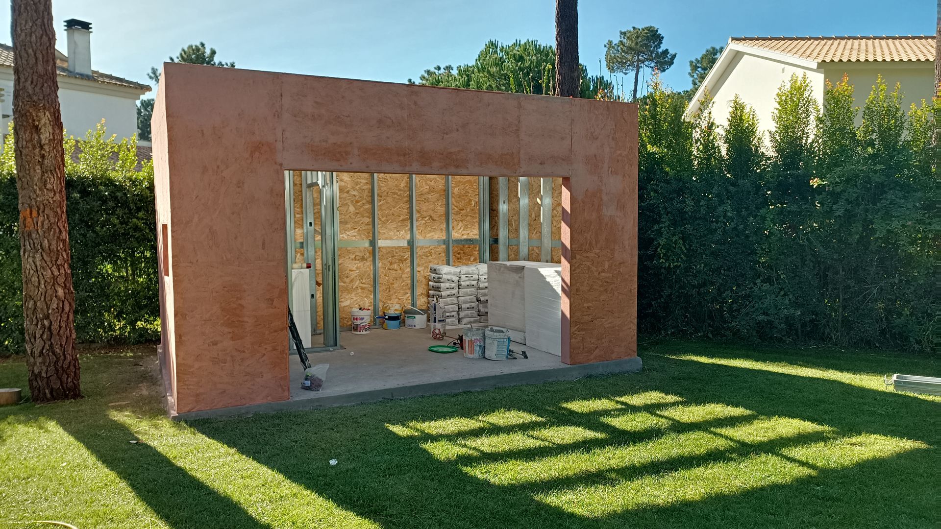 Um pequeno edifício em construção, com paredes vermelho-acastanhadas, interior com estrutura de madeira e um exterior ensolarado.