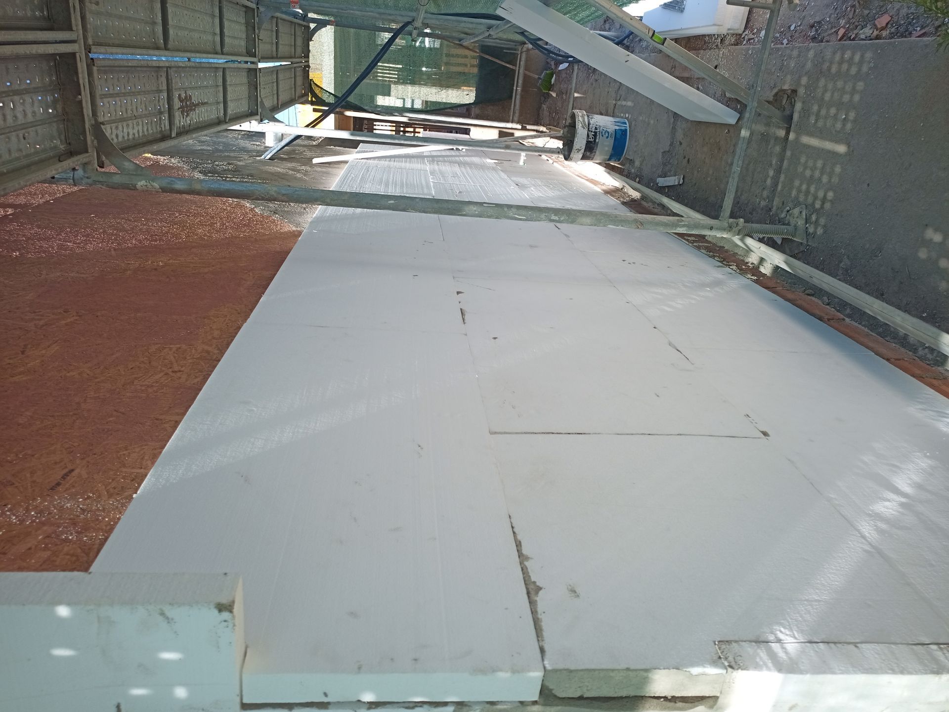 Grandes painéis brancos cobrem uma área de concreto, possivelmente um canteiro de obras, sob um telhado de metal ondulado.