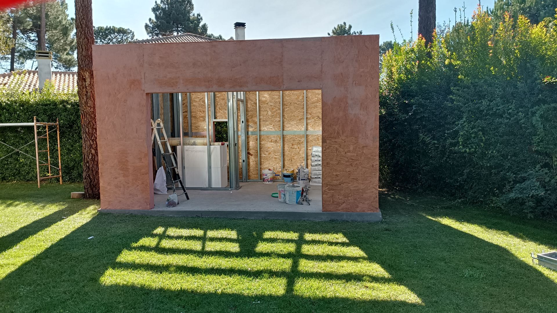 Construção de uma pequena estrutura retangular para uso externo sobre a grama; árvores e uma cerca ao fundo.