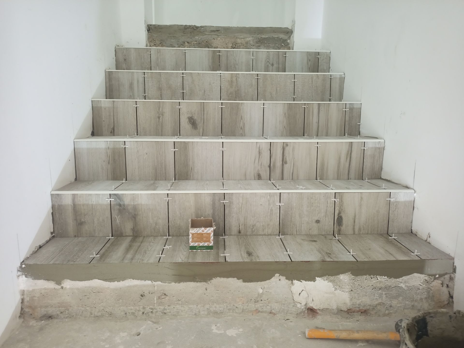 Escada em construção; estão sendo instalados ladrilhos com aparência de madeira nos degraus, com espaçadores já colocados.
