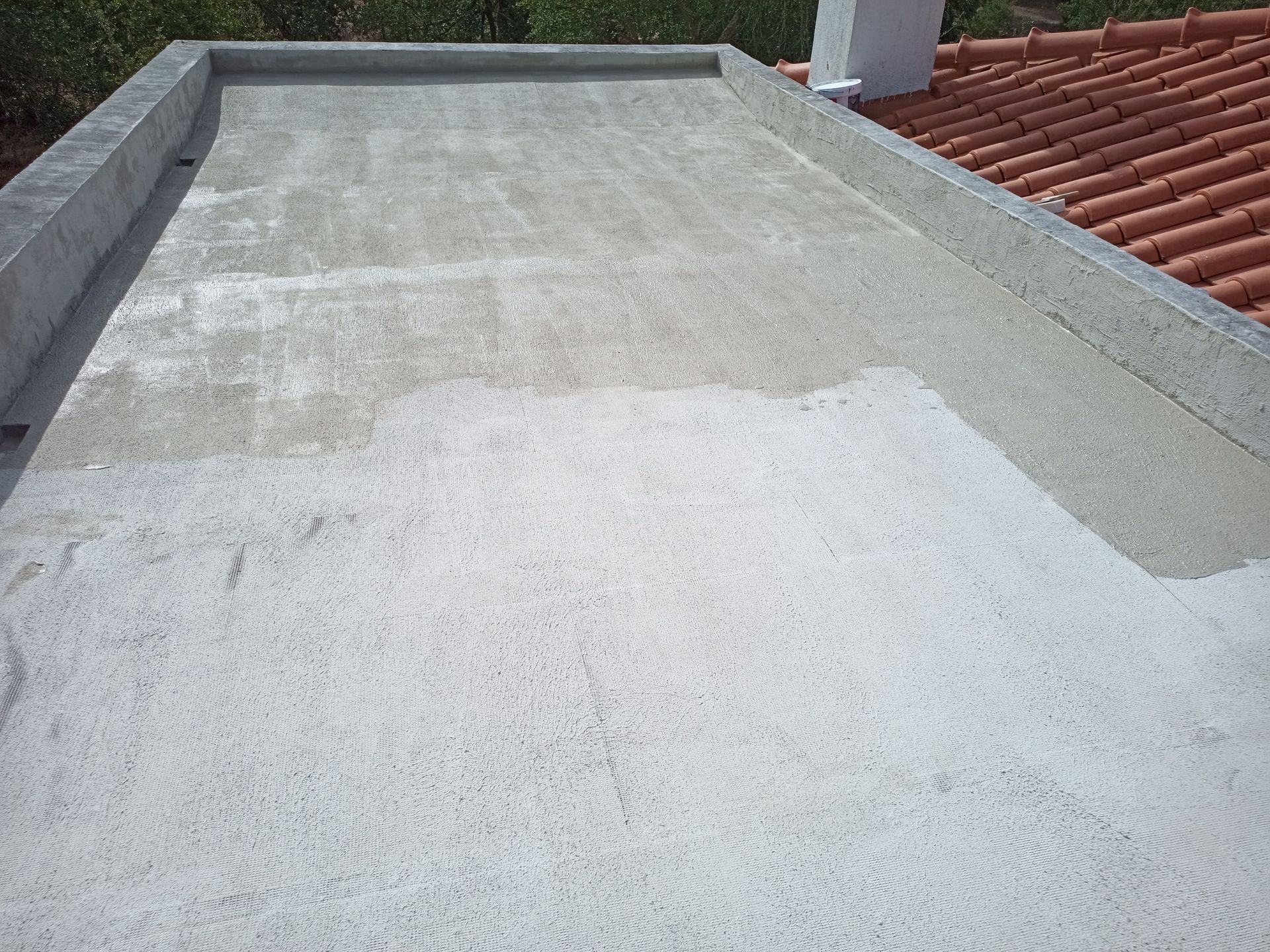 Telhado plano de concreto, parcialmente coberto, ao lado de um telhado de telhas vermelhas e uma chaminé branca.