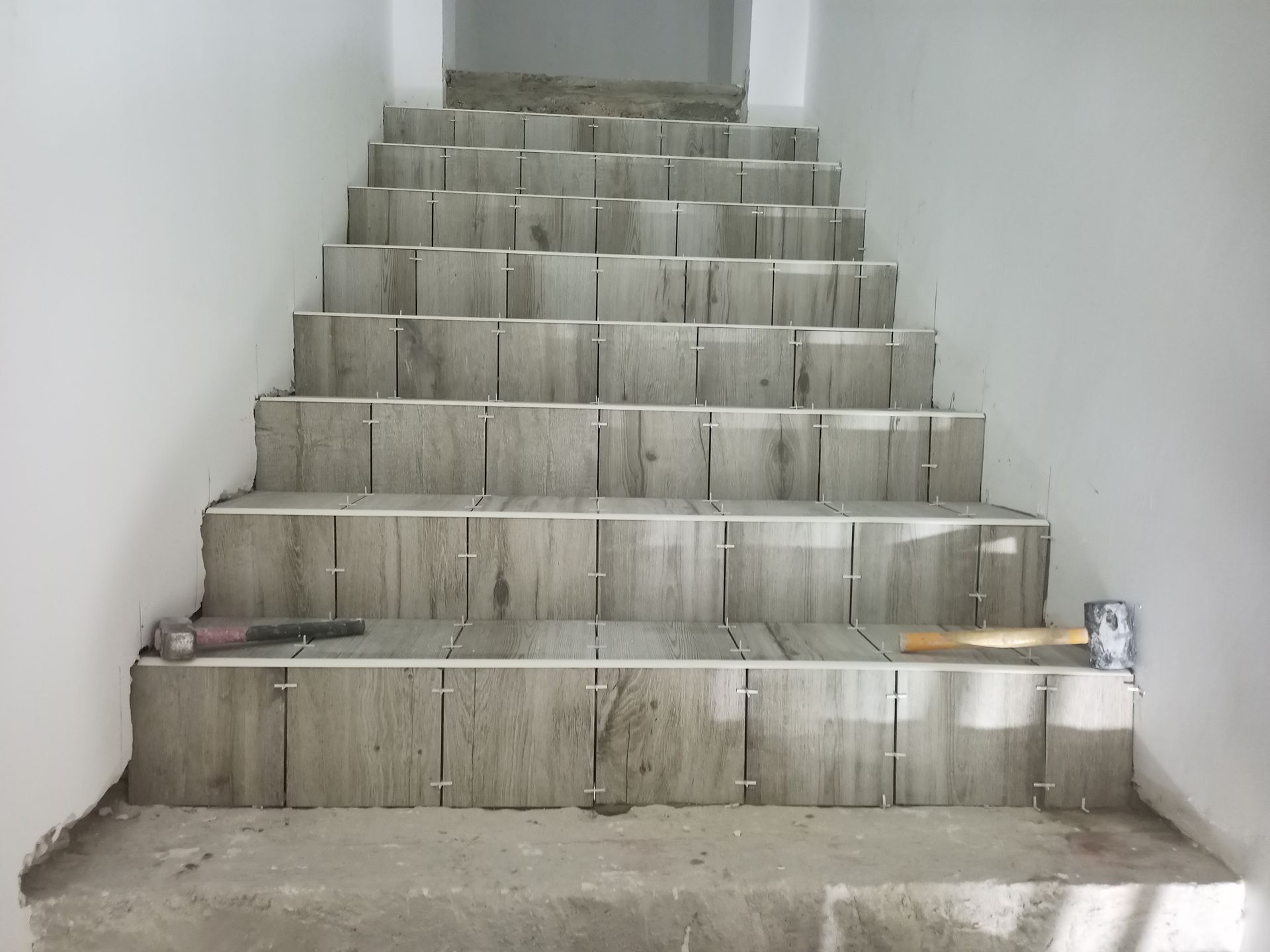 Escada em construção com assentamento de ladrilhos cinza-claro. Ferramentas e espaçadores são visíveis.