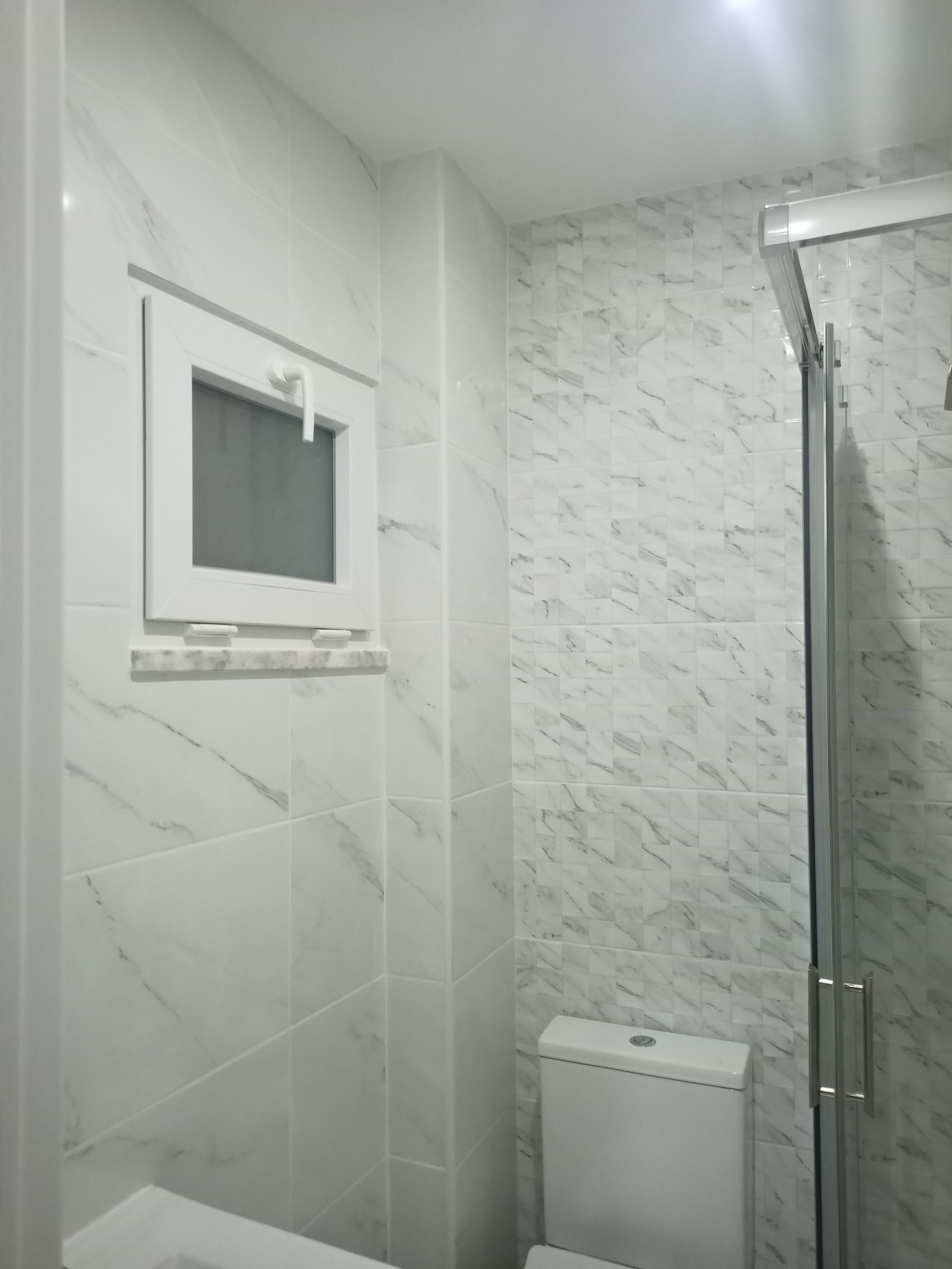 Banheiro branco com azulejos imitando mármore, janela pequena, vaso sanitário e box de vidro.
