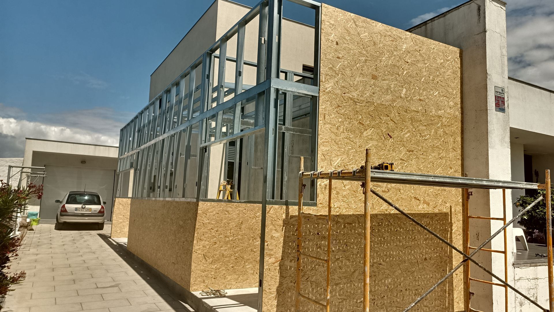 Exterior de um edifício em construção. Estrutura de aço e revestimento de aglomerado visíveis. Andaimes montados. Dia ensolarado.