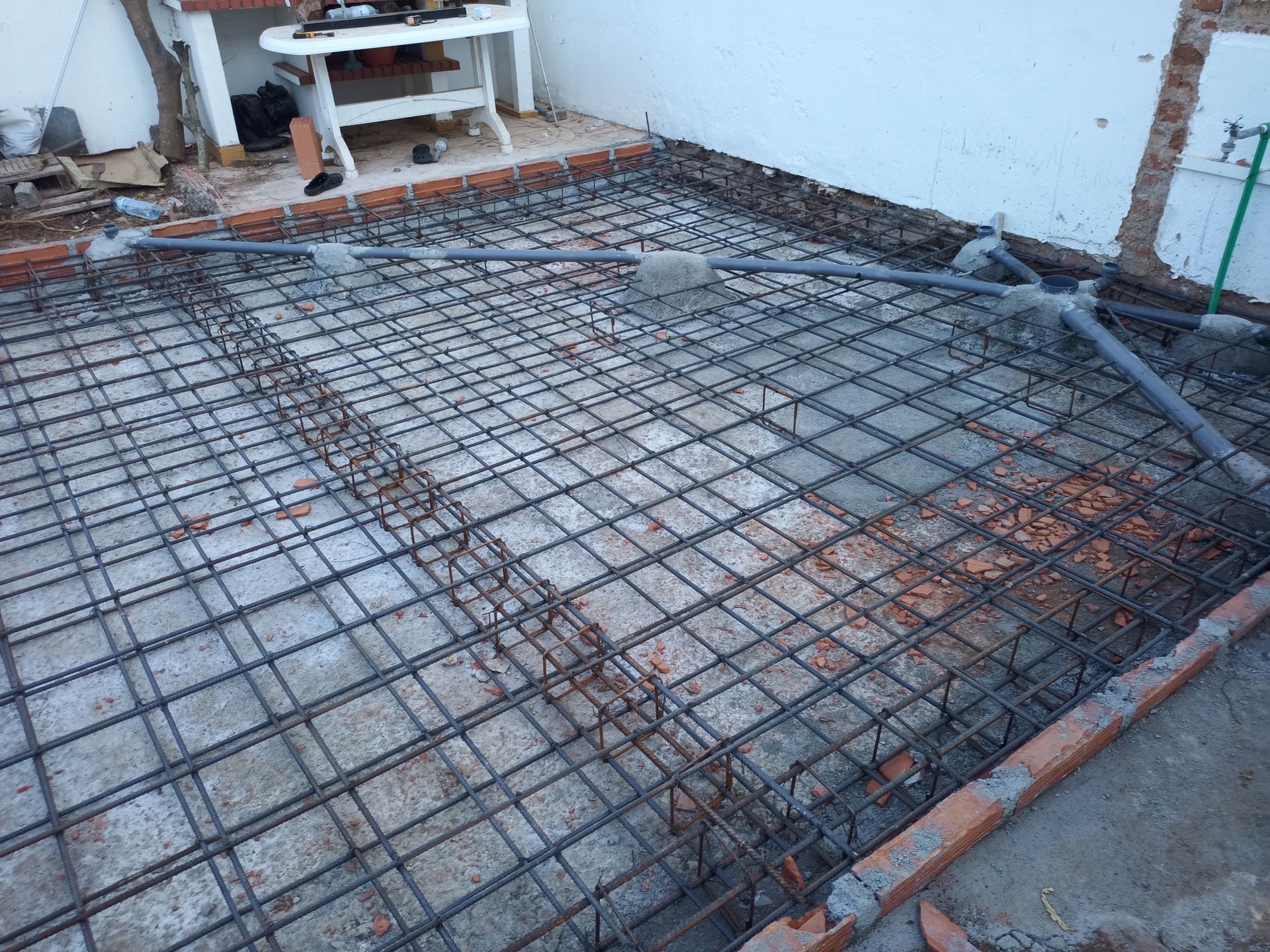 Canteiro de obras: malha de vergalhões de aço, bordas de tijolos e tubulação de PVC sobre fundação de concreto, pronta para a concretagem.