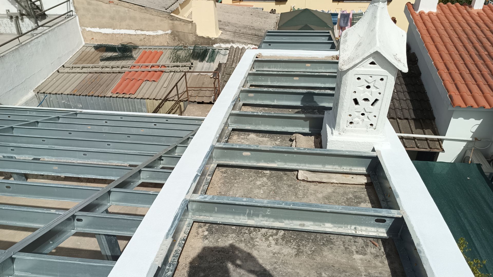 Telhado com estrutura metálica e chaminé, rodeado por outros telhados sob um céu ensolarado.