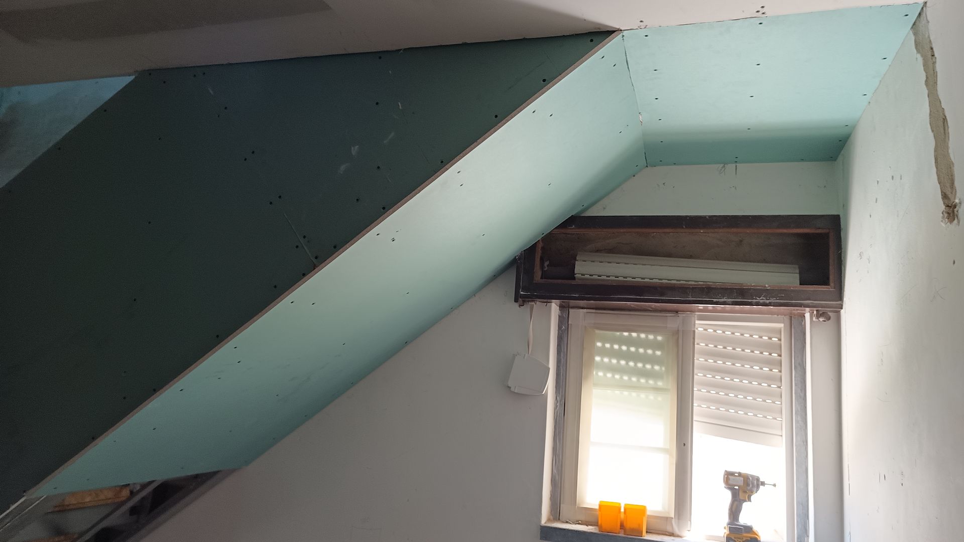 Vista interior com instalação de drywall ao redor de uma janela e área rebaixada, apresentando um tom azul-esverdeado.