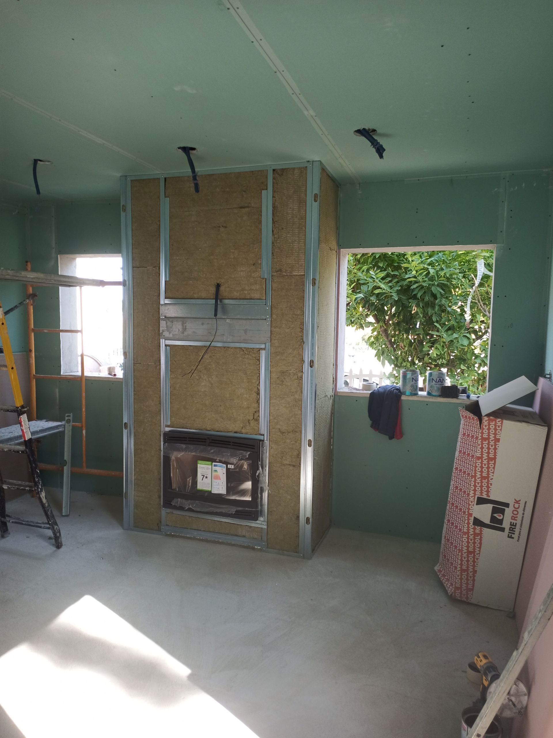Quarto interior em construção, com lareira emoldurada, isolamento e paredes verdes. Duas janelas visíveis.