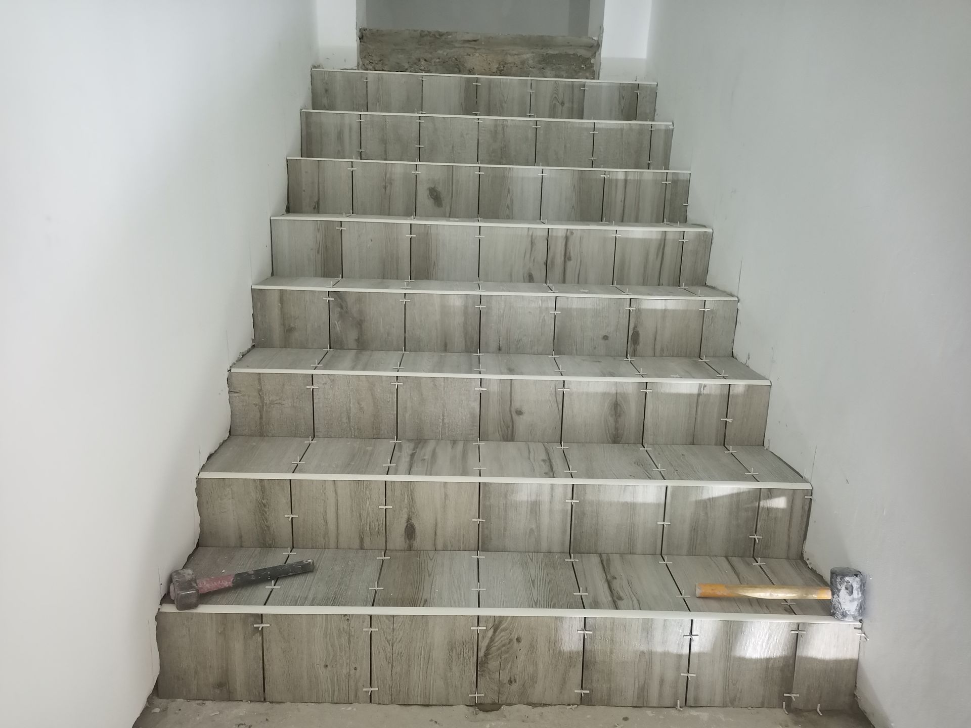 Escada com azulejos cinza-claro sendo instalada. Paredes brancas circundam a escada; ferramentas estão no degrau inferior.