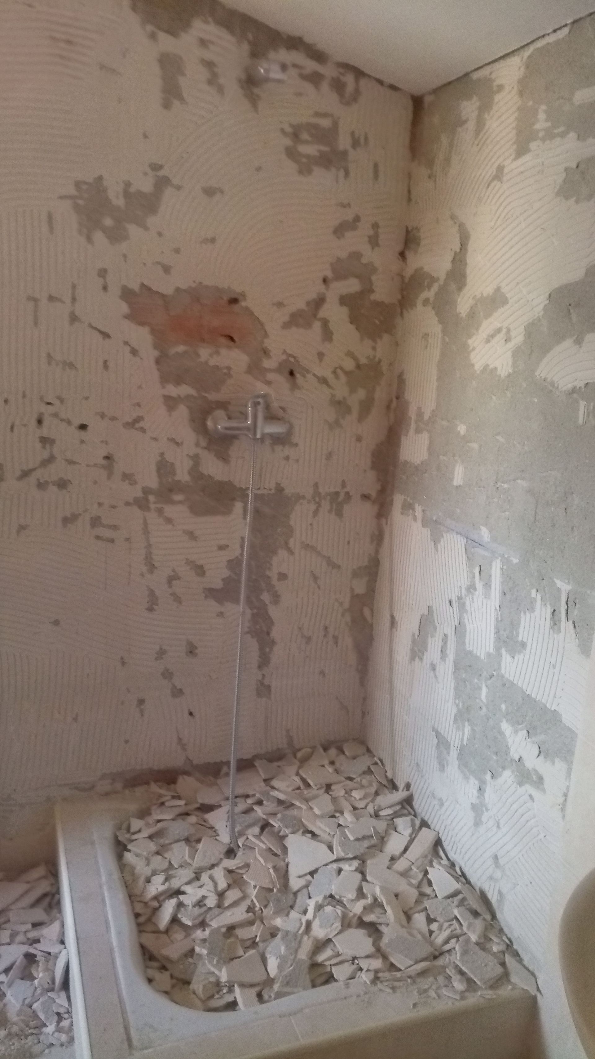 Chuveiro em construção: paredes com reboco descascando, detritos na banheira, chuveiro prateado.