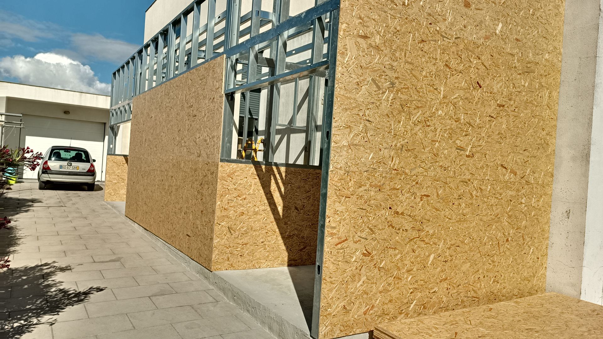 Parede externa em construção: estrutura metálica com painéis de madeira, ao lado de uma entrada de veículos e edifício em concreto.
