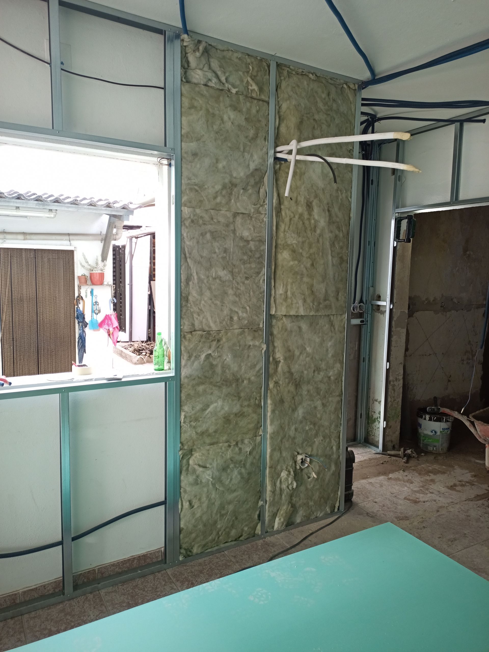 Parede interna em construção com isolamento entre perfis metálicos, próxima a uma janela e porta.